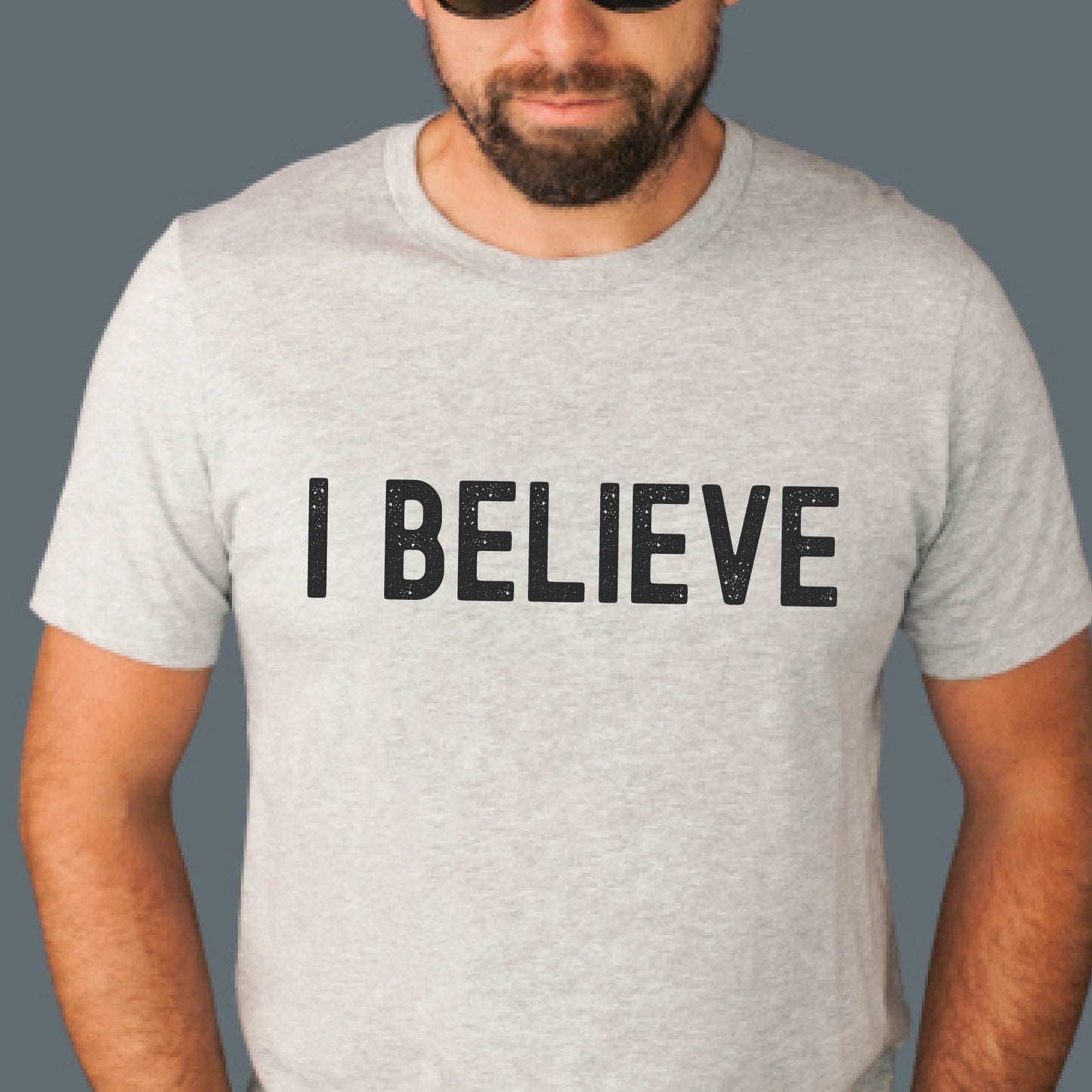 Man or Dad wearing an I BELIEVE Christian aesthetic faith-based bold statement distressed typography t-shirt design printed in matte black on sport gray unisex fit t-shirt, designed for men & women believers