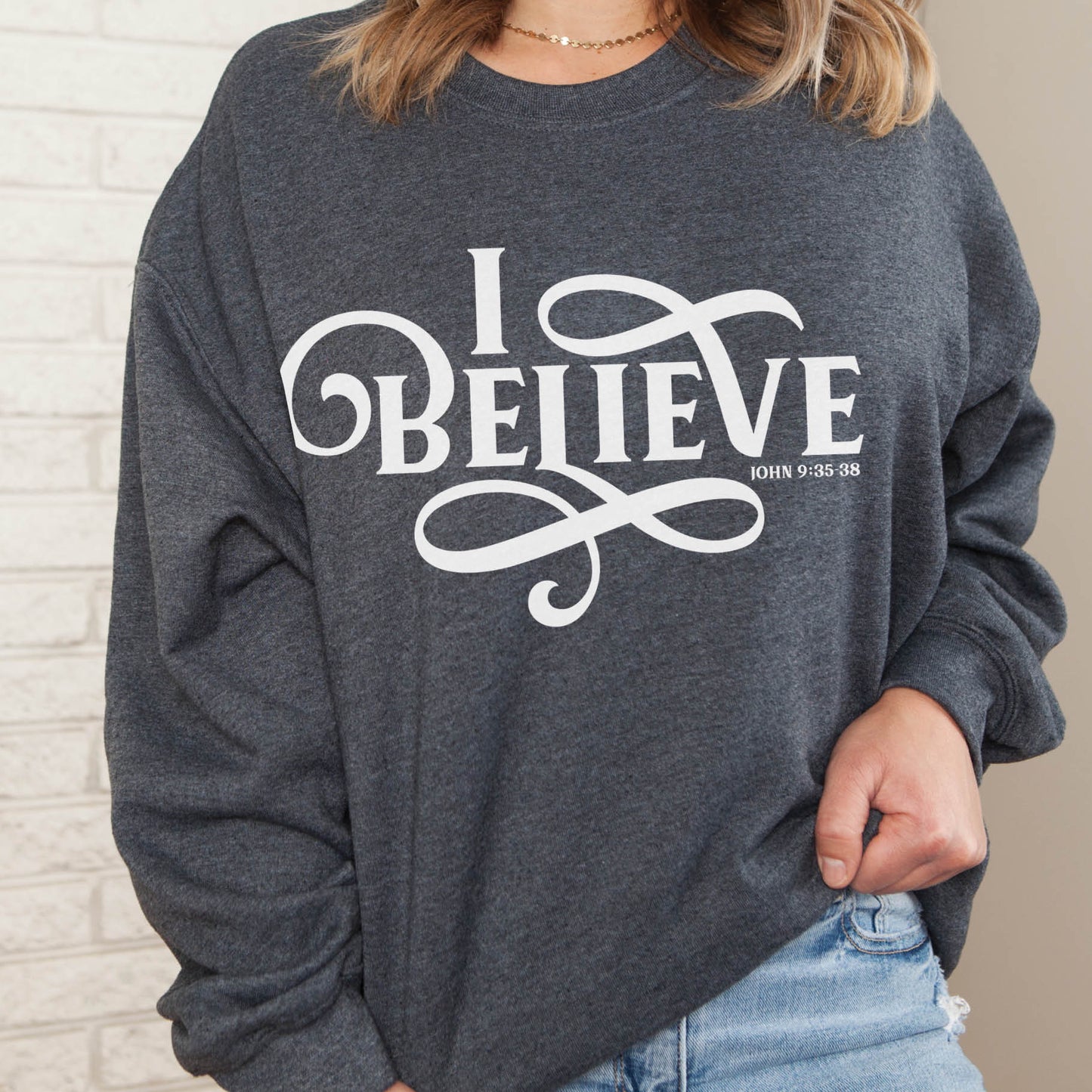 Young woman wearing a heather dark gray I BELIEVE John 9:35-38 bible verse women's Christian unisex cozy sweatshirt, designed for faith-filled Jesus believers in size small, medium, large, XL, 2XL, 3XL, 4XL