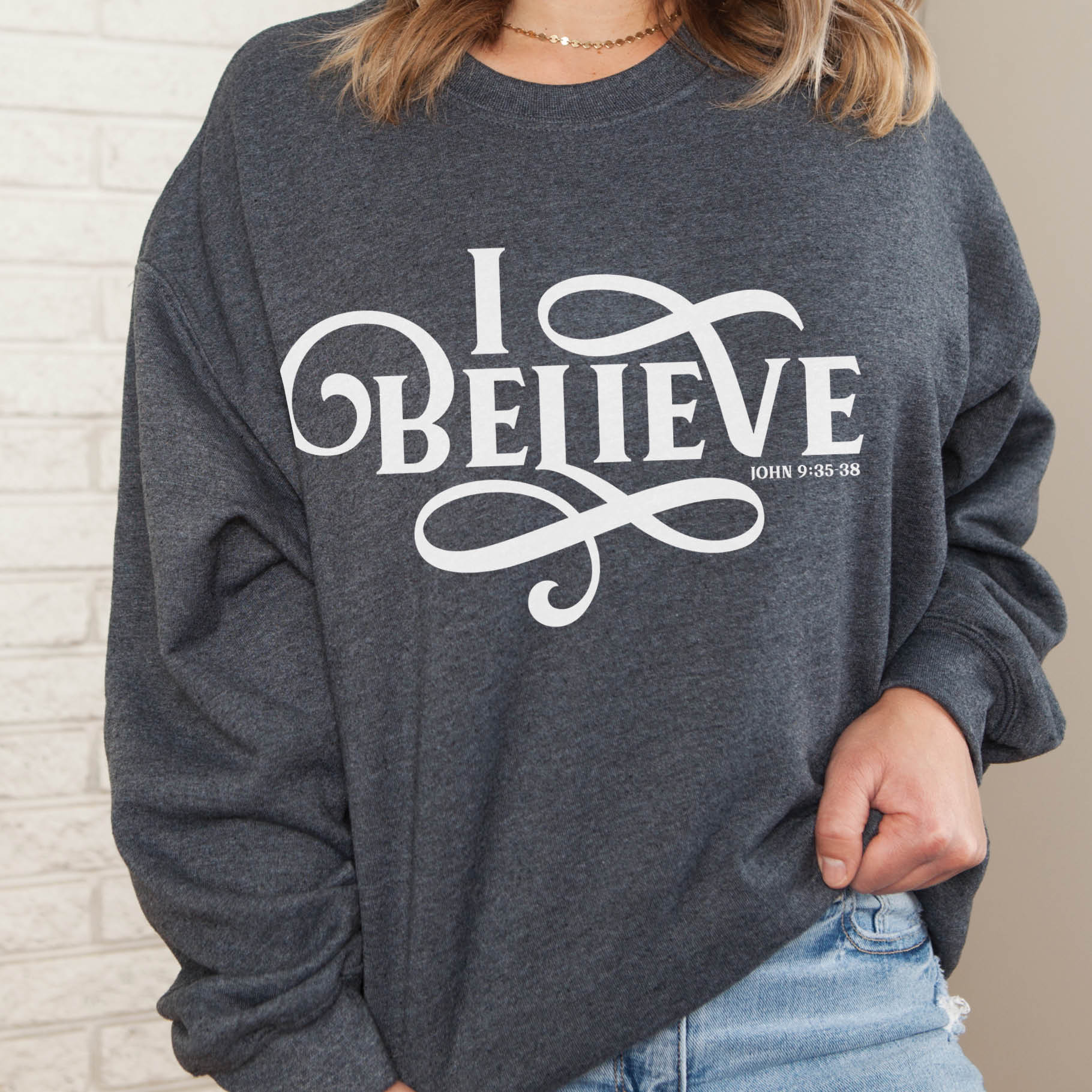 Young woman wearing a heather dark gray I BELIEVE John 9:35-38 bible verse women's Christian unisex cozy sweatshirt, designed for faith-filled Jesus believers in size small, medium, large, XL, 2XL, 3XL, 4XL