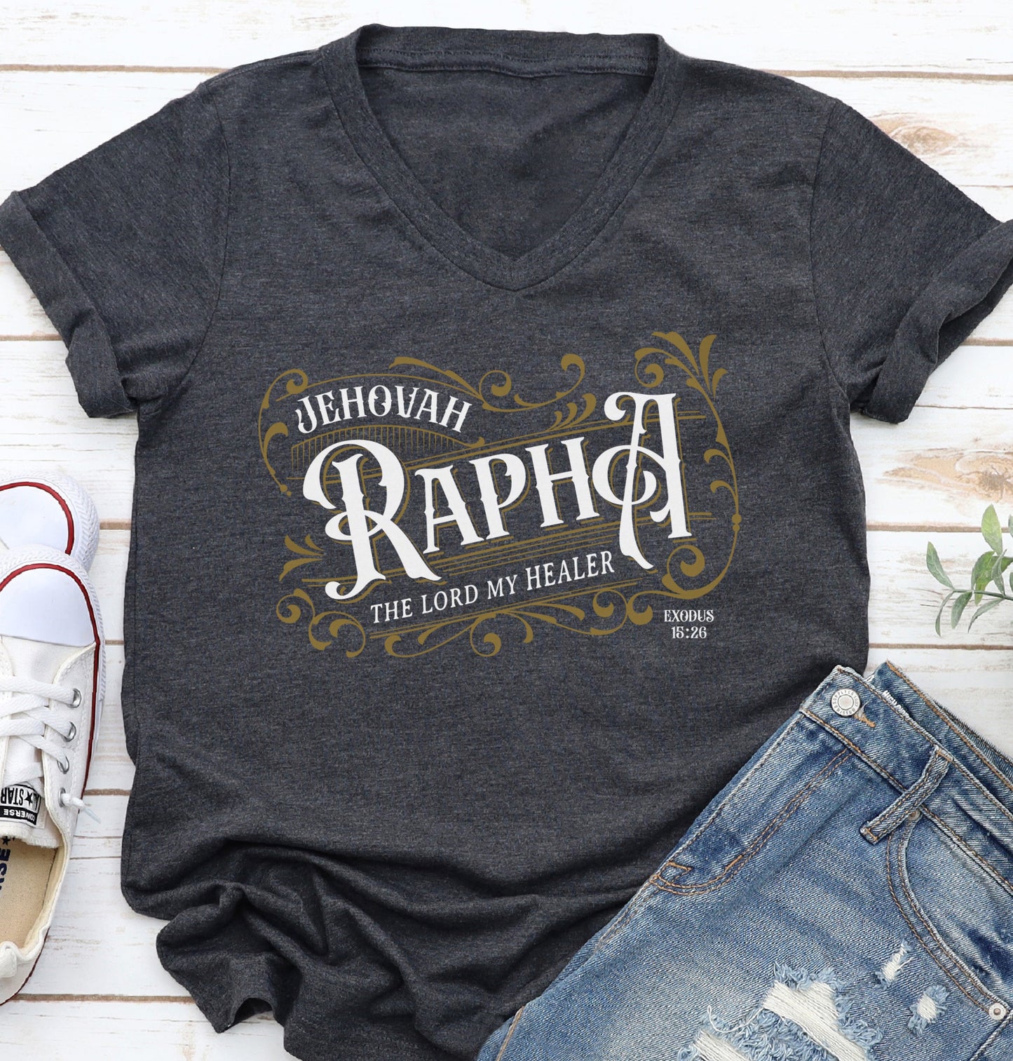 Dark heather gray V-neck women's Christian t-shirt with 'Jehovah Rapha, The Lord My Healer, Exodus 18:26 bible verse' vintage blackletter style design laying flat on a white wooden surface with jeans and sneakers