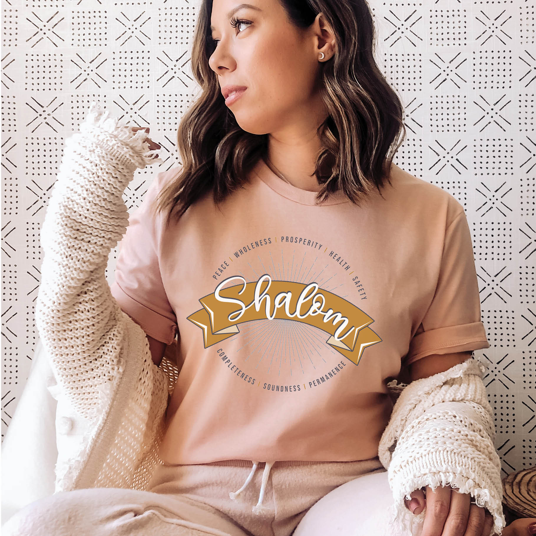 Young woman wearing a soft heather prism peach faith-based Christian women's unisex t-shirt with a gold banner and sunburst design that says the Biblical Hebrew meaning of the word "SHALOM" - Peace, Completeness, Prosperity, Health, Safety, Wholeness, Soundness, and Permanence definition, makes a great mother's day gift for mom!