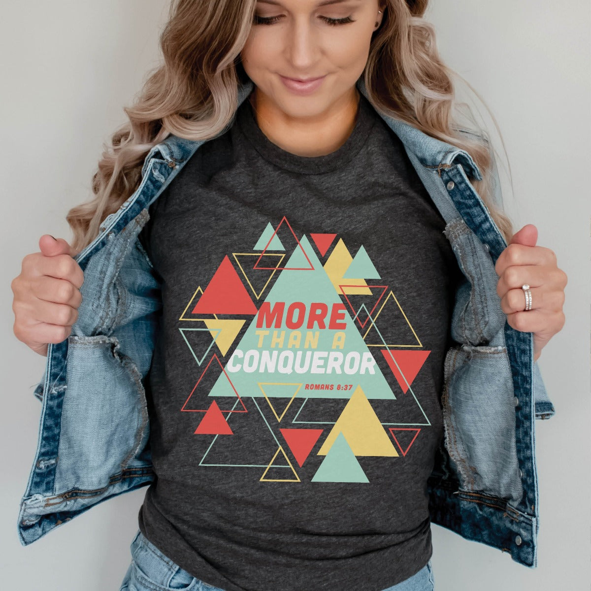 Modern young woman wearing a heather dark gray "More Than A Conqueror" Romans 8:37 bible verse scripture unisex faith-based Christian Bella Canvas 3001 t-shirt, with bright and colorful geometric triangles pattern, made in the USA for Men and Women Jesus believers