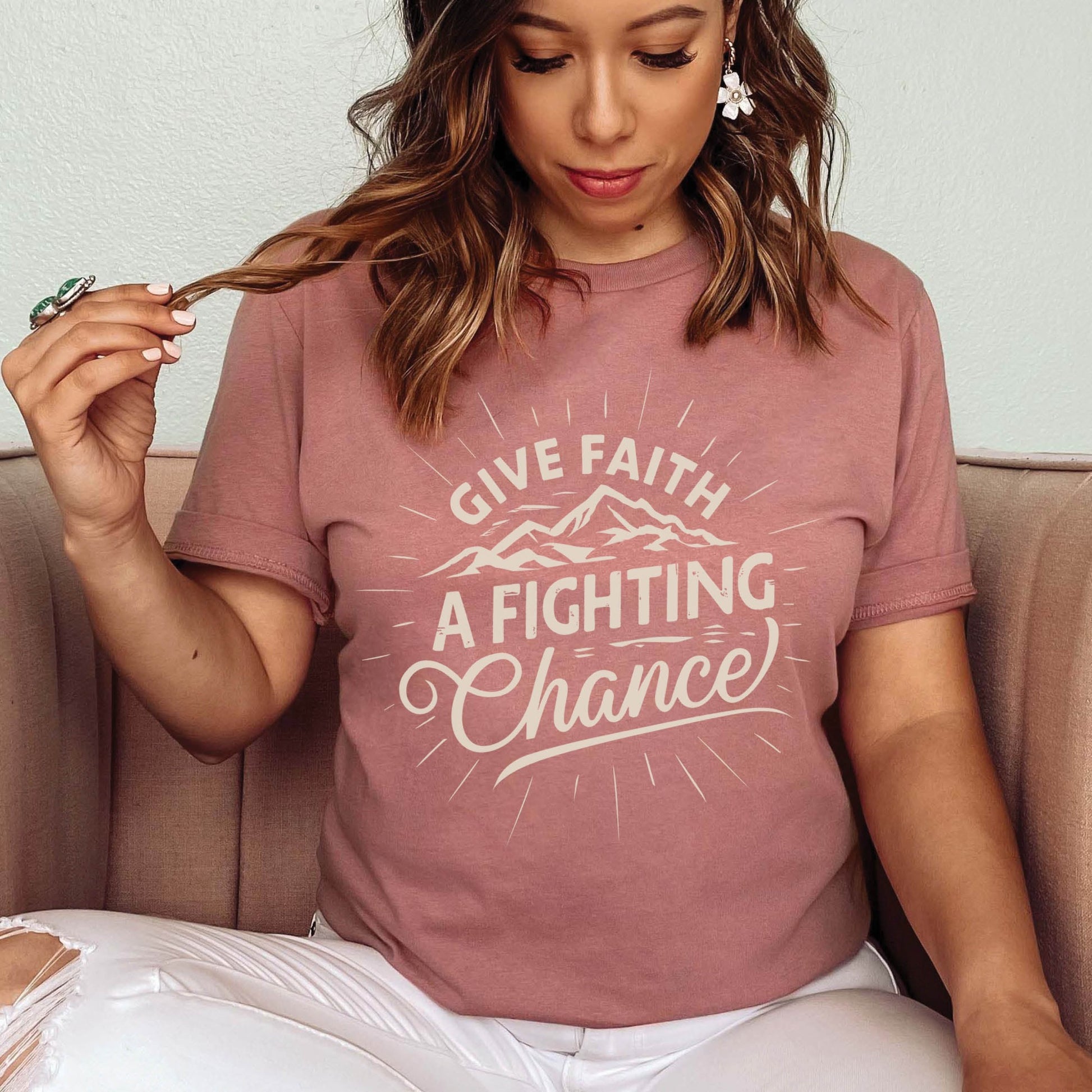Modern young woman wearing a mauve pink Unisex Tee with Christian Bible verse quote that says, "Give Faith A Fighting Chance" with retro sunburst and mountain graphics, t-shirt designed for faith-based men and women