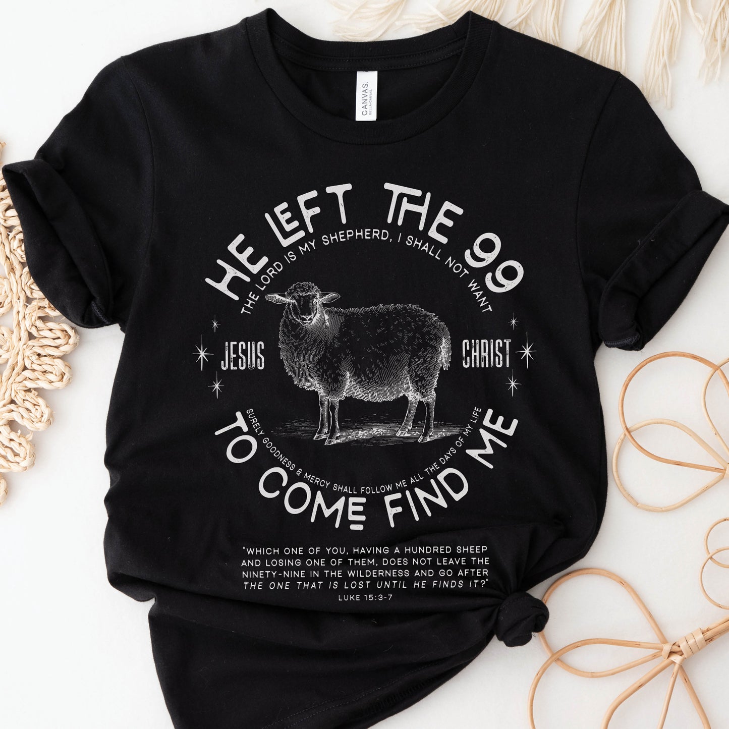 Black Christian unisex t-shirt laying on woven raffia mats with He Left the 99 to Come Find Me Jesus Christ Luke 15 bible verse and vintage sheep illustration graphic and distressed text printed in white