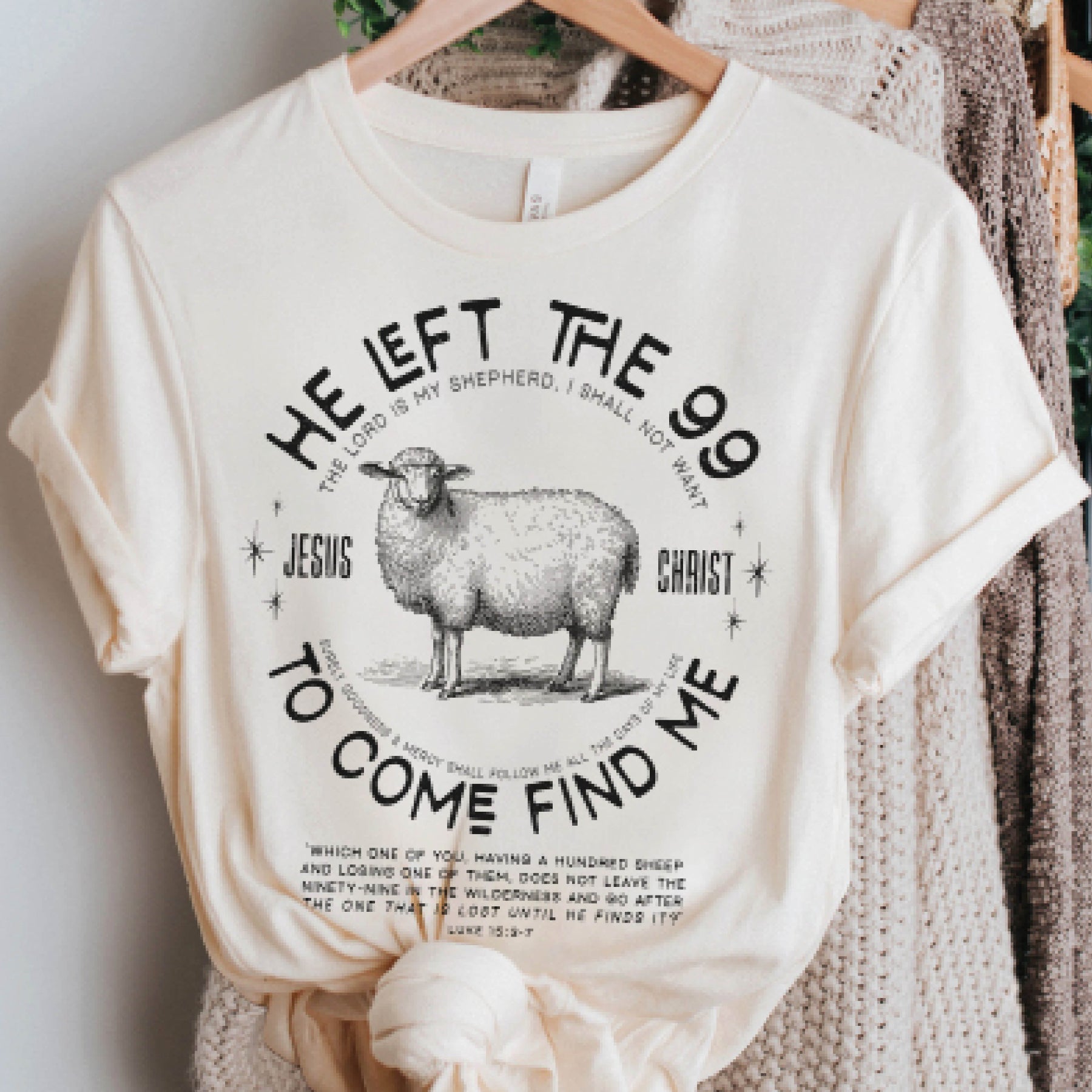 Soft cream Christian unisex t-shirt hanging on a wooden hanger with cozy sweaters behind it with He Left the 99 to Come Find Me Jesus Christ Luke 15 bible verse and vintage sheep illustration graphic and distressed text printed in black