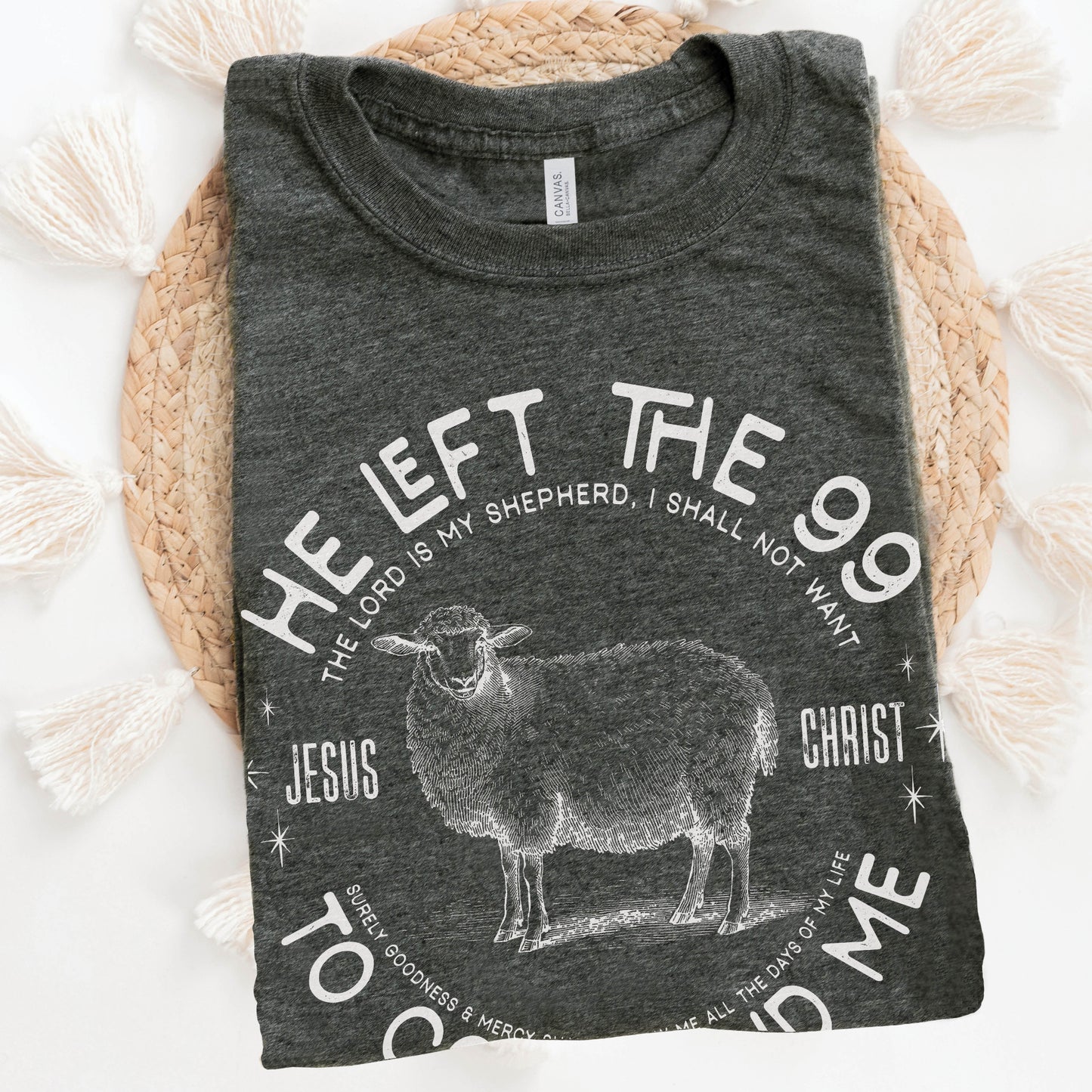 Folded heather dark gray Christian unisex t-shirt laying on a woven raffia mat with He Left the 99 to Come Find Me Jesus Christ Luke 15 bible verse and vintage sheep illustration graphic and distressed text printed in white