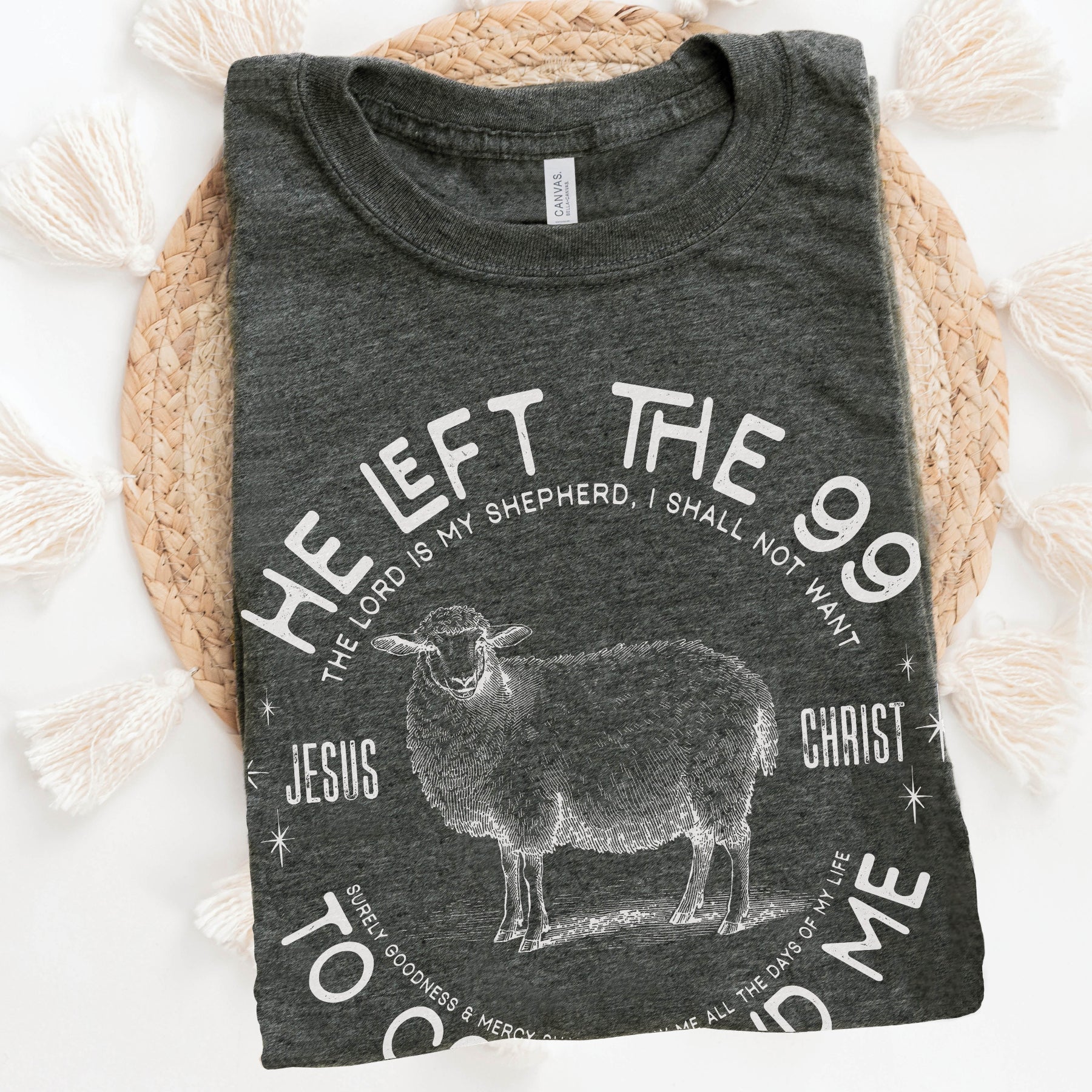 Folded heather dark gray Christian unisex t-shirt laying on a woven raffia mat with He Left the 99 to Come Find Me Jesus Christ Luke 15 bible verse and vintage sheep illustration graphic and distressed text printed in white