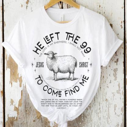 White Christian unisex t-shirt laying on a on a wooden background with He Left the 99 to Come Find Me Jesus Christ Luke 15 bible verse and vintage sheep illustration graphic and distressed text printed in black