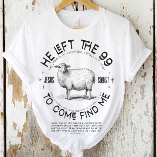 White Christian unisex t-shirt laying on a on a wooden background with He Left the 99 to Come Find Me Jesus Christ Luke 15 bible verse and vintage sheep illustration graphic and distressed text printed in black