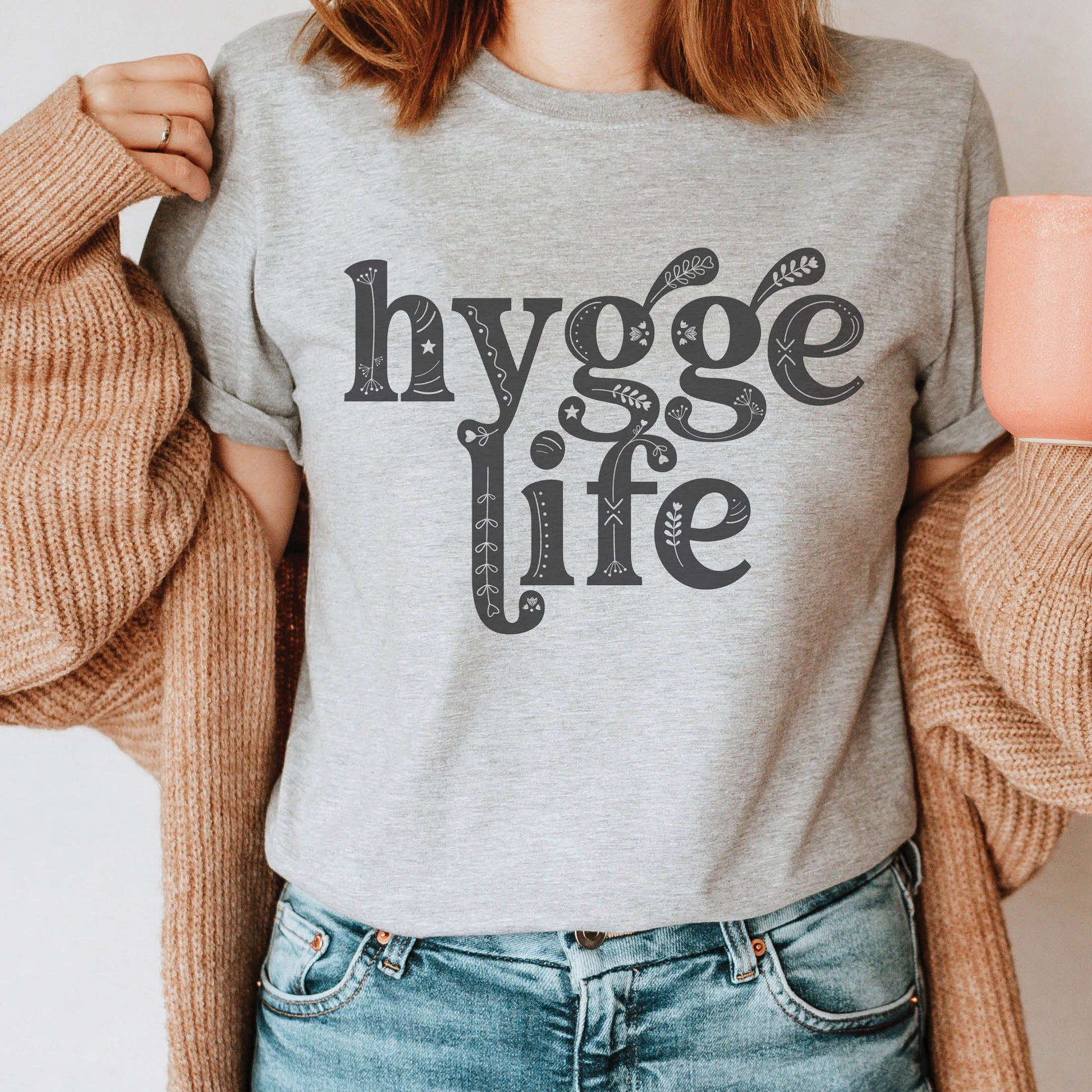 Coziness vibes woman wearing a heather gray Hygge Life Holy Hygge Women's unisex Christian t-shirt with Scandinavian floral and heart art for the cozy fall and winter season