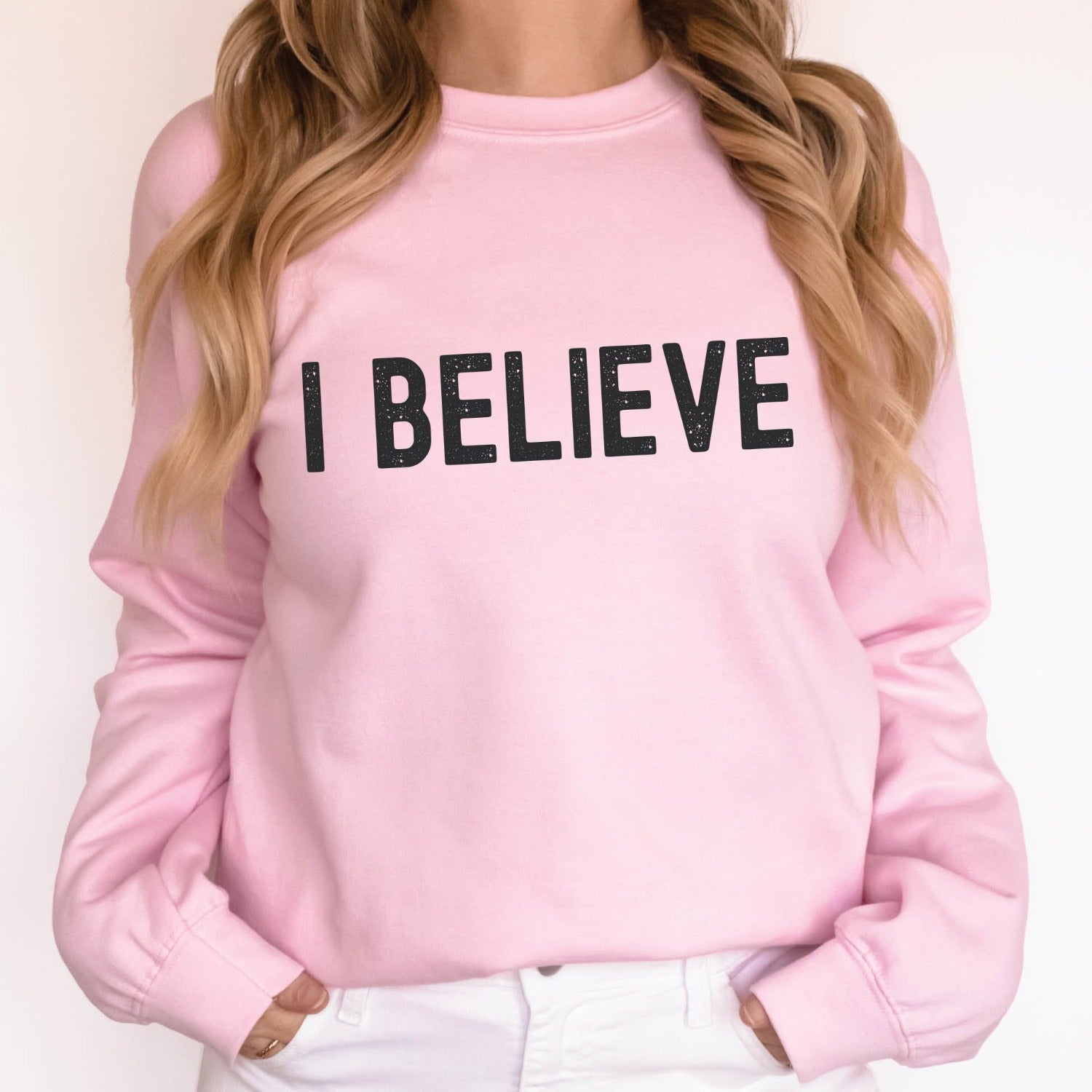 Woman wearing an "I Believe" Christian aesthetic Jesus believer matte black simple distressed typography design printed on cozy light pink unisex crewneck sweatshirt, created for men & women