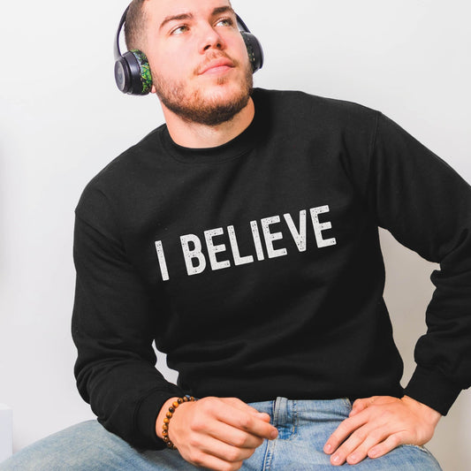 Young man with headphones wearing an "I Believe" Christian aesthetic Jesus believer simple distressed white typography design printed on cozy black unisex crewneck sweatshirt, created for men & women