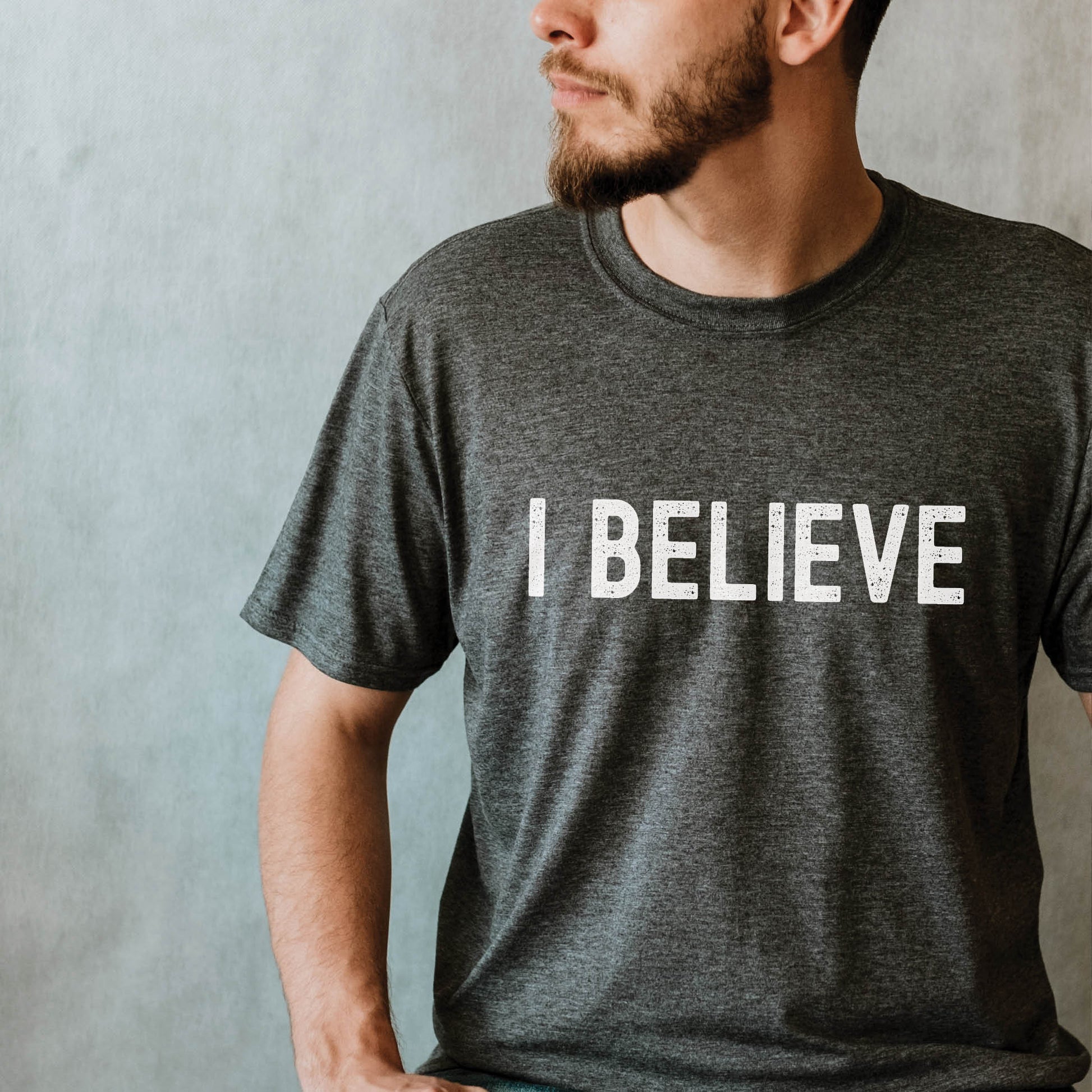 Young man wearing an I BELIEVE Christian aesthetic faith-based bold statement distressed typography t-shirt design printed in white on a heather dark gray unisex fit t-shirt, designed for men & women believers