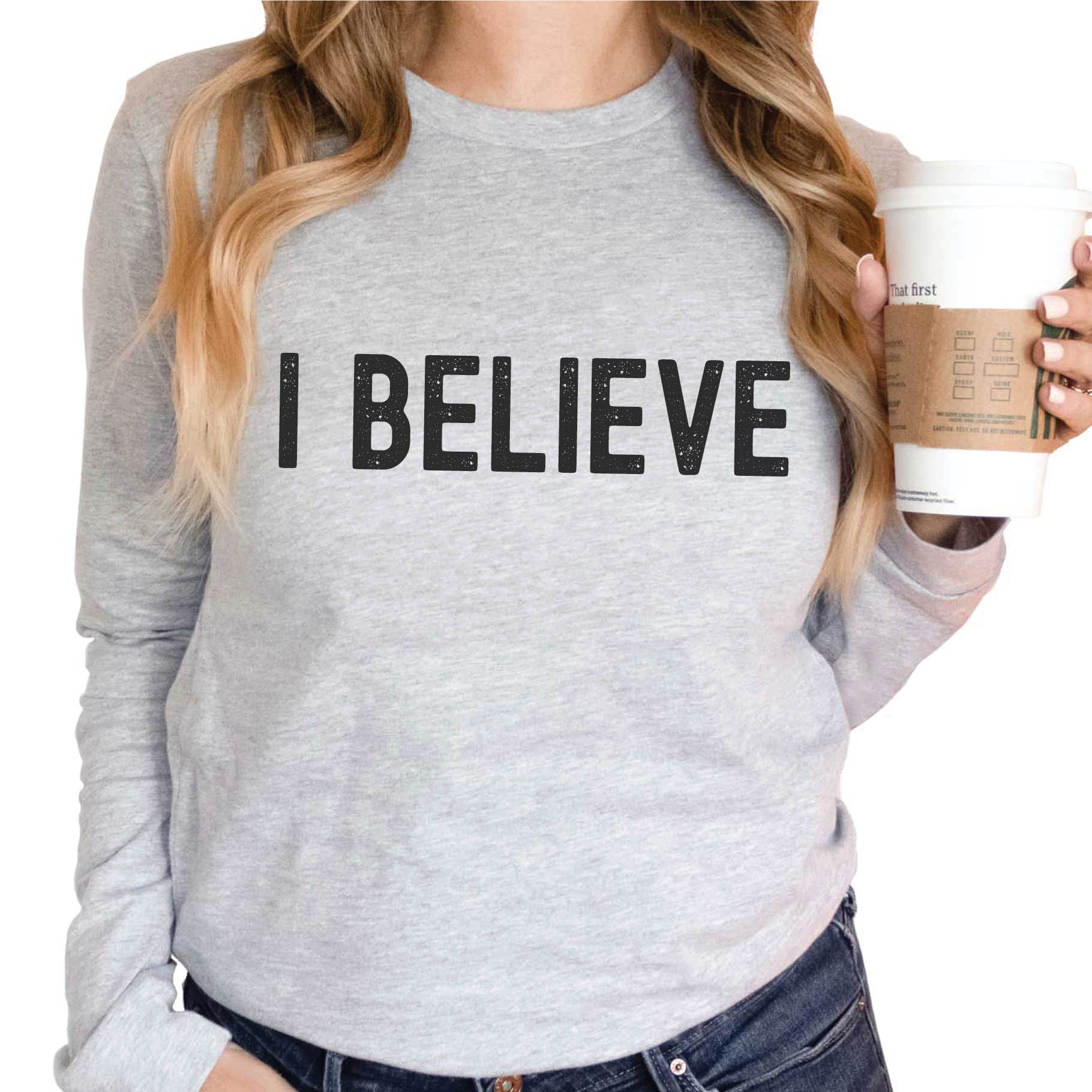 Coffee drinking woman wearing a heather gray cozy long sleeve t-shirt with a bold faith-based statement "I BELIEVE" distressed typography design printed in black, created for Christian men and women Jesus believers