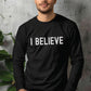 Man wearing a black cozy long sleeve t-shirt with a bold faith-based statement "I BELIEVE" distressed typography design printed in white, created for Christian men and women Jesus believers