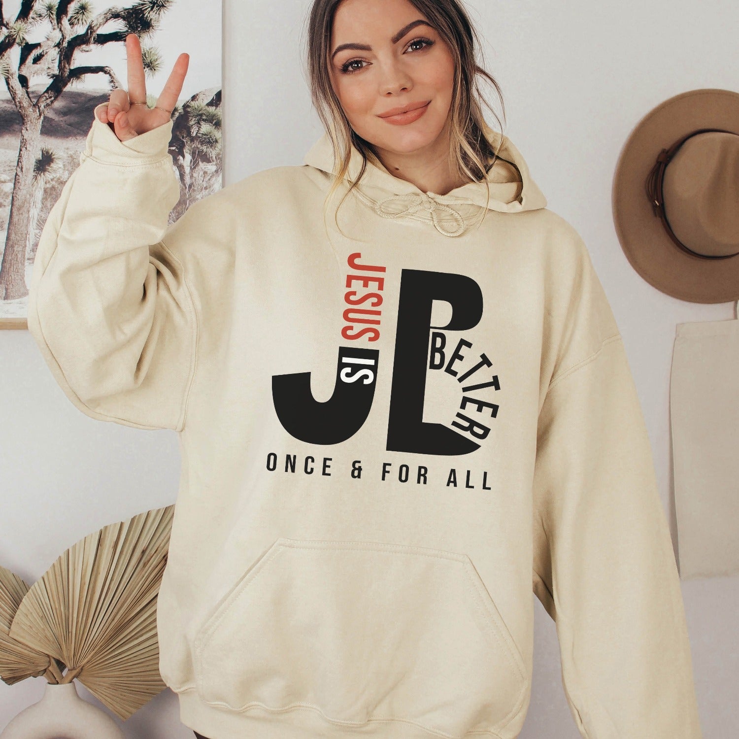 Woman like a mom wearing a cozy "JB" typography "Jesus is Better Once and For All" design in black and red letters, based on the Christian bible book of Hebrews, printed on a sand ivory color unisex hoodie, created for faith-based men and women, great mother's day gift for her
