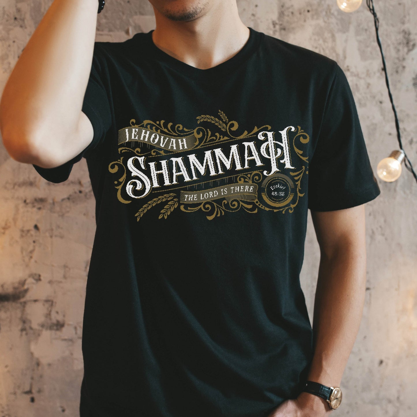 Young man wearing a gold Jehovah Shammah "The Lord Is There" Christian unisex black t-shirt from Ezekiel 38:35 vision bible verse Hebrew meaning, with vintage blackletter and ornate decorative swirl filigree design, created for men and women believers