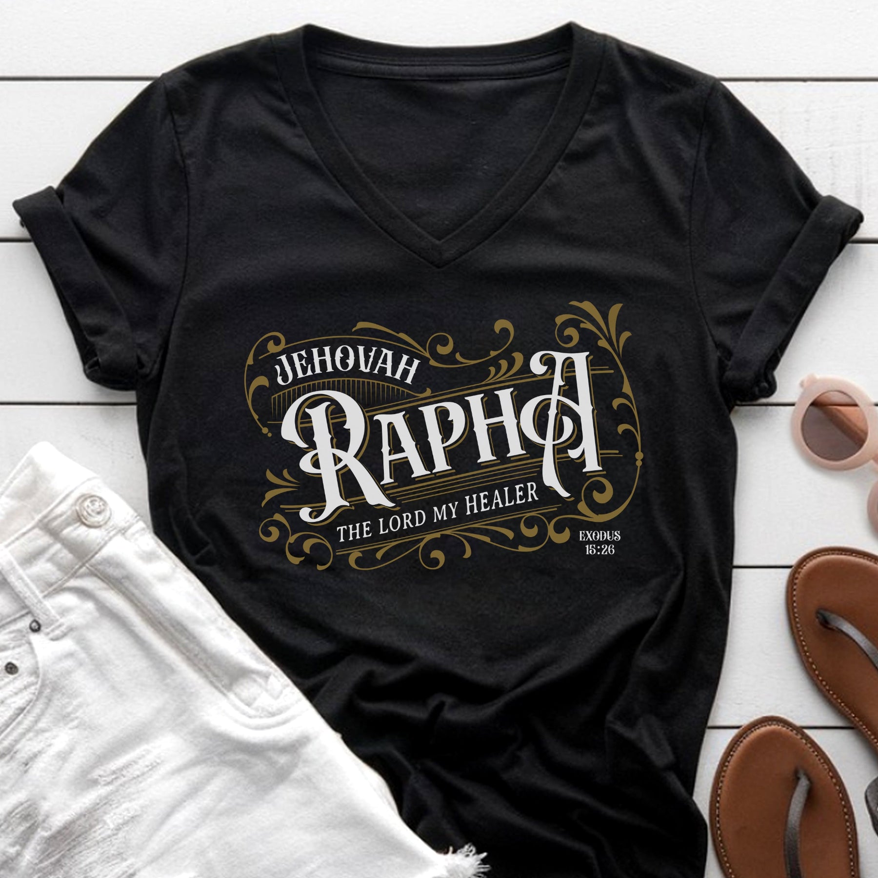 Black V-neck women's Christian t-shirt with 'Jehovah Rapha' vintage blackletter design on a white wooden surface with sunglasses and sandals.