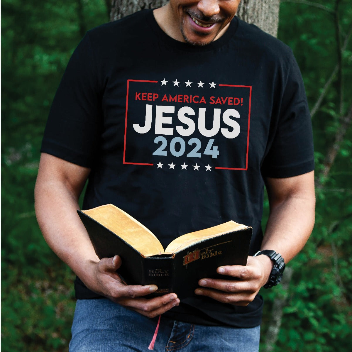 Patriotic man reading the bible wearing black Bella Canvas 3001 Presidential election vote unisex Christian t-shirt with "Jesus 2024 Keep America Saved" printed in bold red, white, and blue fonts, tees made in the USA for men and women God & Country Patriots