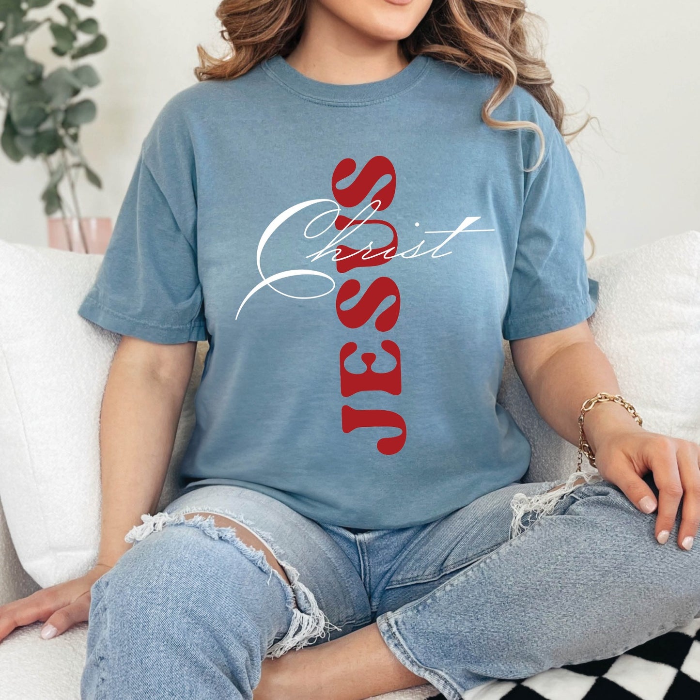 Young woman wearing a dusty ice blue color Christian Comfort Colors 1717 heavyweight unisex t-shirt with faith-based bible typography that says, "Jesus Christ" and white and red letters in the shape of the cross, created for men and women Kingdom believers