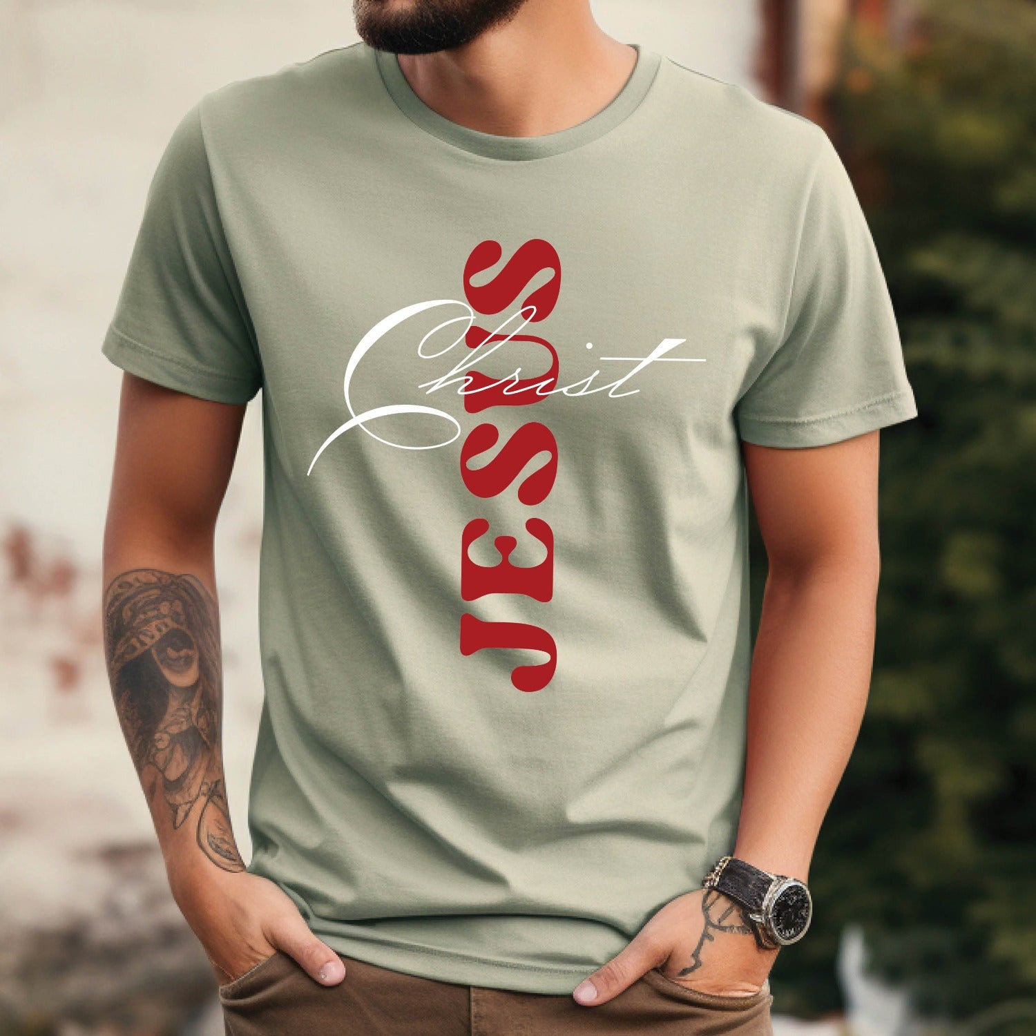 Christian man wearing a Bay sage green color Comfort Colors 1717 heavyweight unisex t-shirt with faith-based bible typography that says, "Jesus Christ" and white and red letters in the shape of the cross, created for men and women Kingdom believers