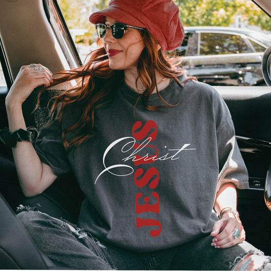 Trendy young woman wearing an oversized Pepper dark gray color Christian unisex Comfort Colors 1717 heavyweight t-shirt with faith-based bible typography that says, "Jesus Christ" and white and red letters in the shape of the cross, created for men and women Kingdom believers
