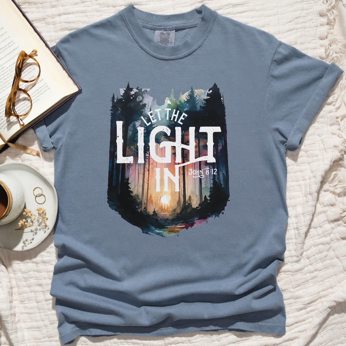 Blue Jean dusty blue color garment dyed Comfort Colors C1717 unisex faith-based Christian t-shirt with "Let the Light In" John 8:12 bible verse and watercolor moody forest trees scene, designed for men and women