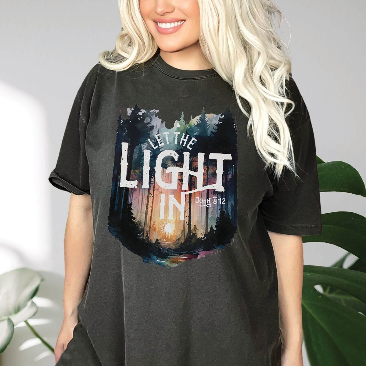 Beautiful young lady wearing trendy oversized Pepper distressed black Comfort Colors C1717 unisex faith-based Christian t-shirt with "Let the Light In" John 8:12 bible verse and watercolor moody forest trees scene, designed for men and women
