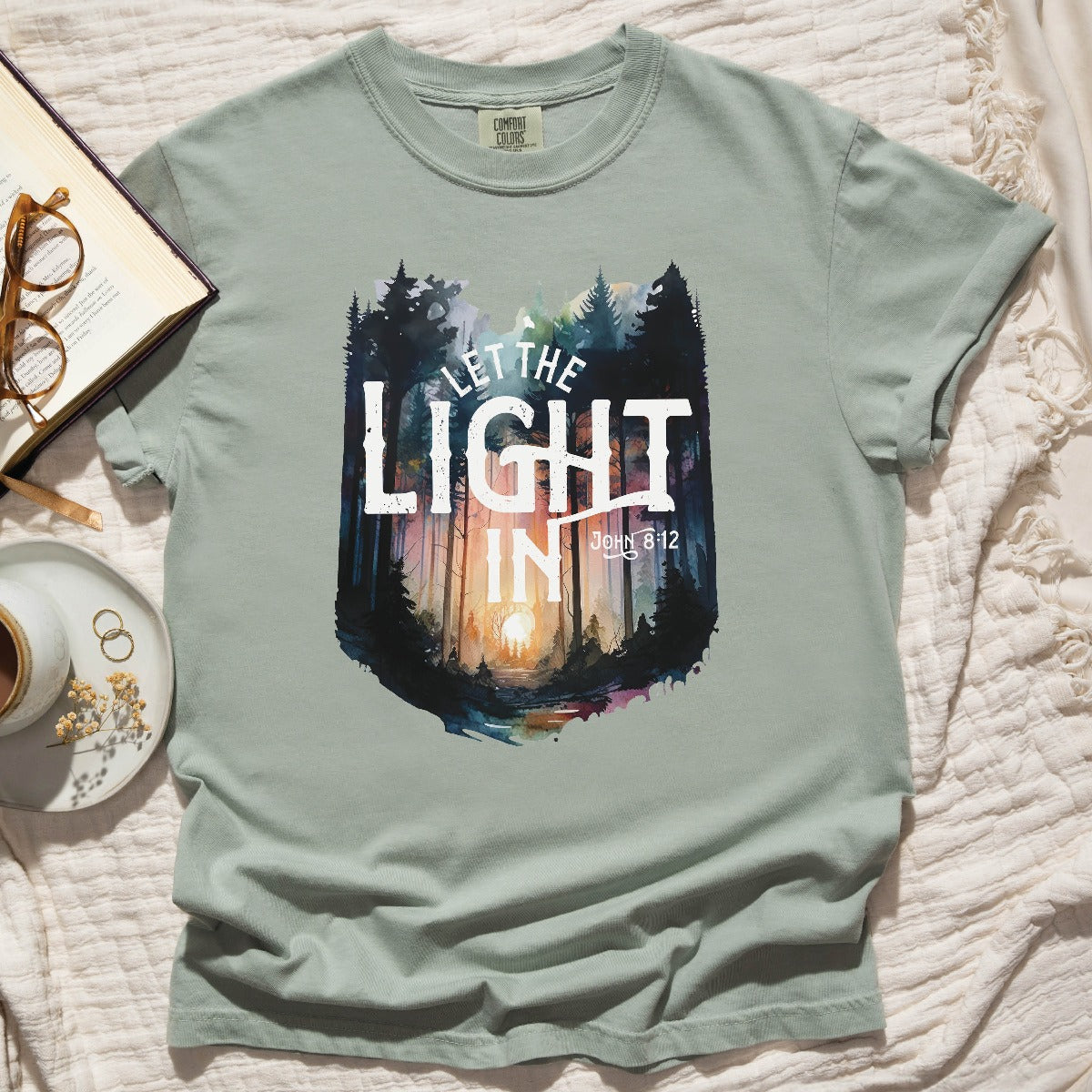 Bay light blue / green garment dyed Comfort Colors C1717 unisex faith-based Christian t-shirt with "Let the Light In" John 8:12 bible verse and watercolor moody forest trees scene, designed for men and women