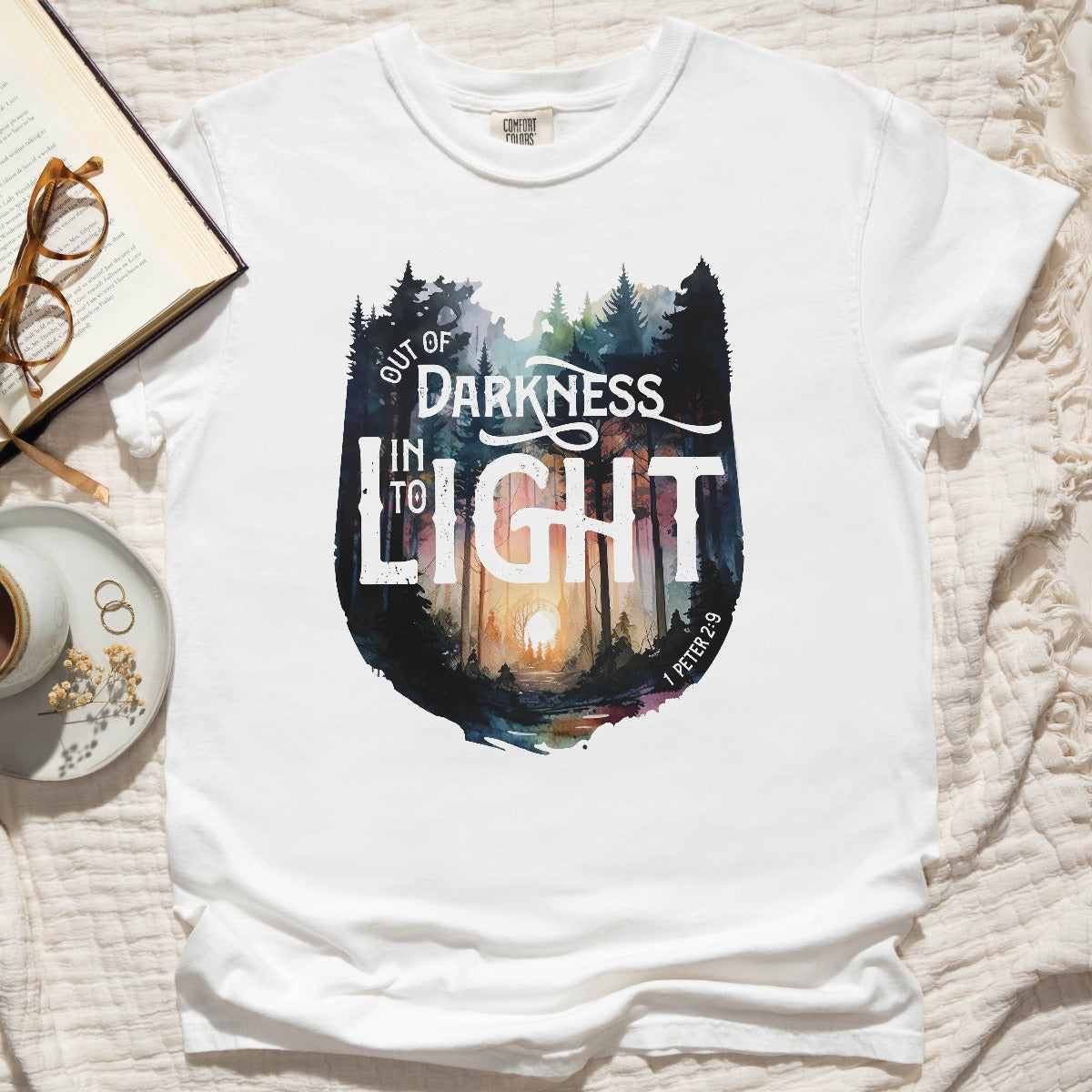 White Comfort Colors C1717 unisex faith-based Christian t-shirt with "Out of Darkness, Into Light" 1 Peter 2:9 bible verse and watercolor moody forest trees outdoor scene, designed for men and women