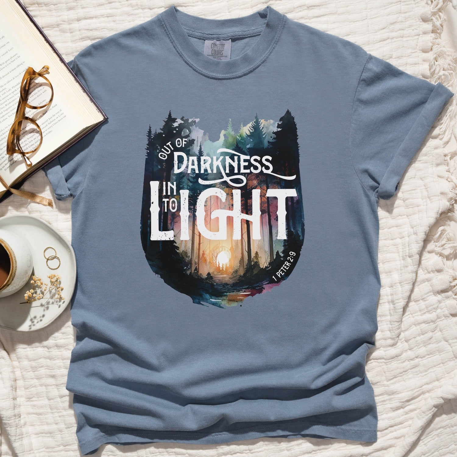 Blue Jean dusty blue color garment dyed Comfort Colors C1717 unisex faith-based Christian t-shirt with "Out of Darkness, Into Light" 1 Peter 2:9 bible verse and watercolor moody forest trees outdoor scene, designed for men and women