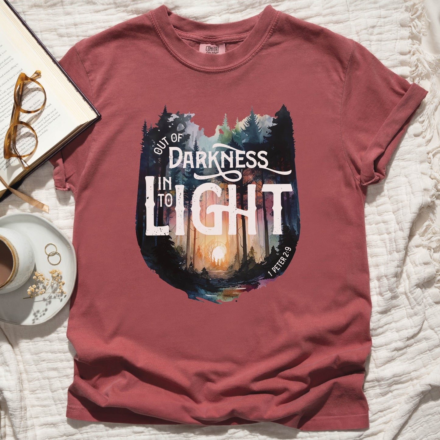 Crimson light dusty red color garment dyed Comfort Colors C1717 unisex faith-based Christian t-shirt with "Out of Darkness, Into Light" 1 Peter 2:9 bible verse and watercolor moody forest trees outdoor scene, designed for men and women