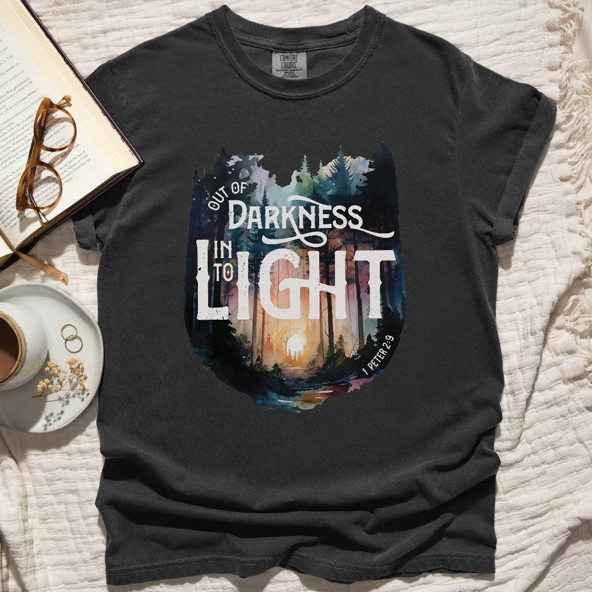 Pepper distressed black color garment dyed Comfort Colors C1717 unisex faith-based Christian t-shirt with "Out of Darkness, Into Light" 1 Peter 2:9 bible verse and watercolor moody forest trees outdoor scene, designed for men and women