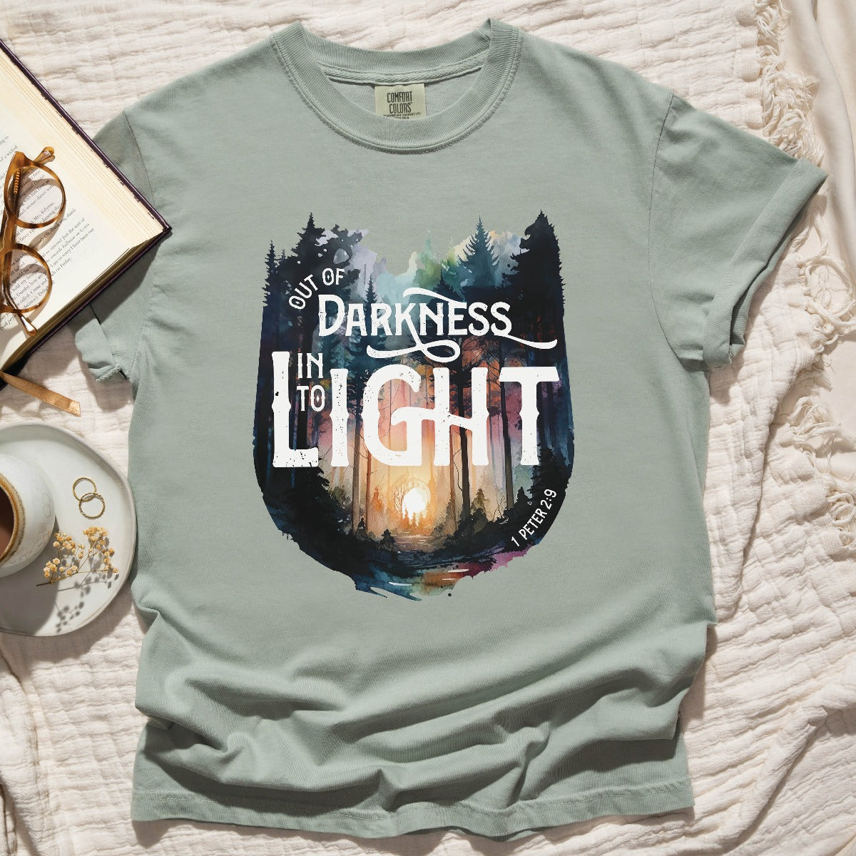 Bay light sage green color garment dyed Comfort Colors C1717 unisex faith-based Christian t-shirt with "Out of Darkness, Into Light" 1 Peter 2:9 bible verse and watercolor moody forest trees outdoor scene, designed for men and women