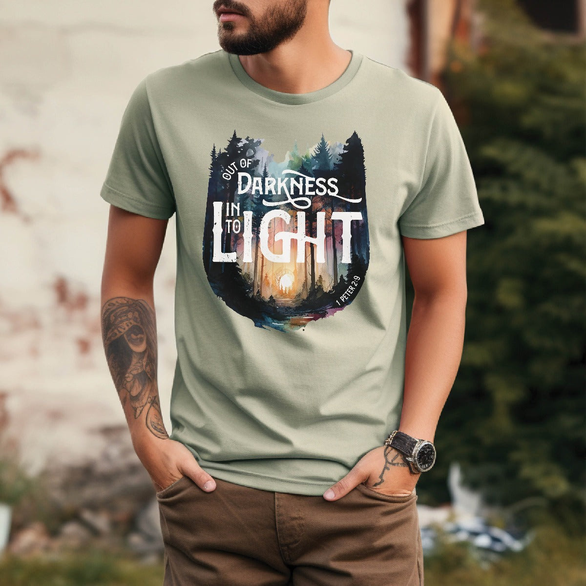 Young trendy man wearing Bay light sage green color garment dyed Comfort Colors C1717 unisex faith-based Christian t-shirt with "Out of Darkness, Into Light" 1 Peter 2:9 bible verse and watercolor moody forest trees outdoor scene, designed for men and women
