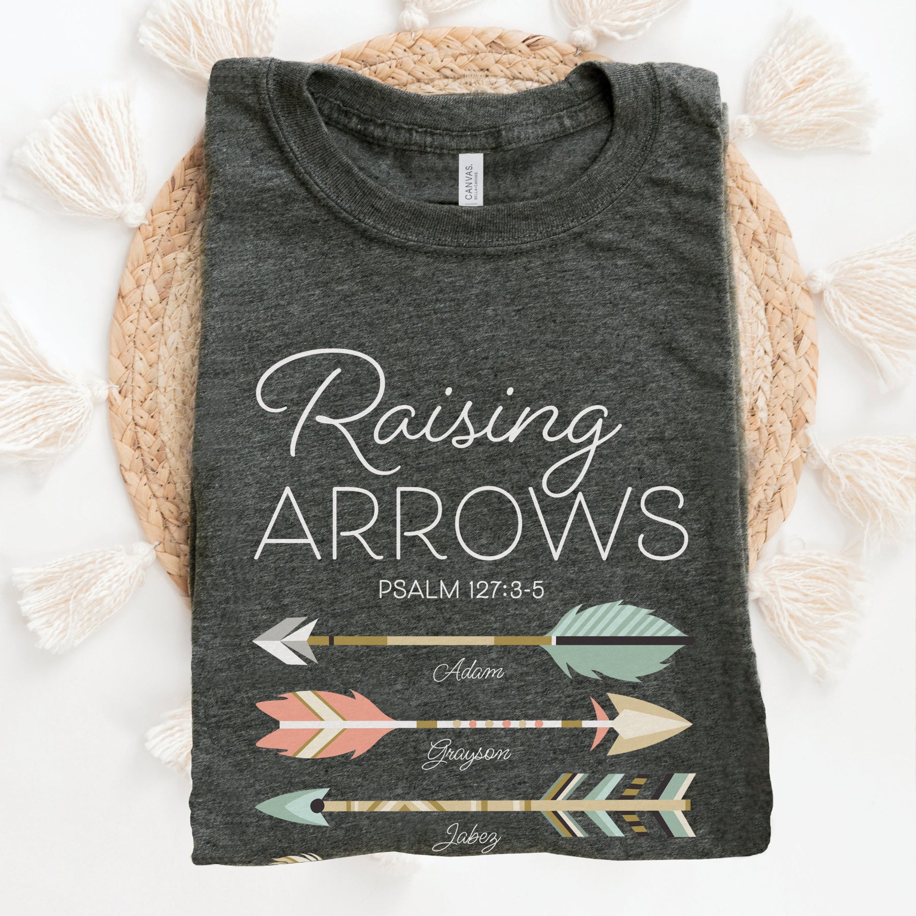 A folded dark heather gray t-shirt with 'Raising Arrows' Psalm 127:3-5 design with 5 personalized names under each arrow and boho accessories on a white background