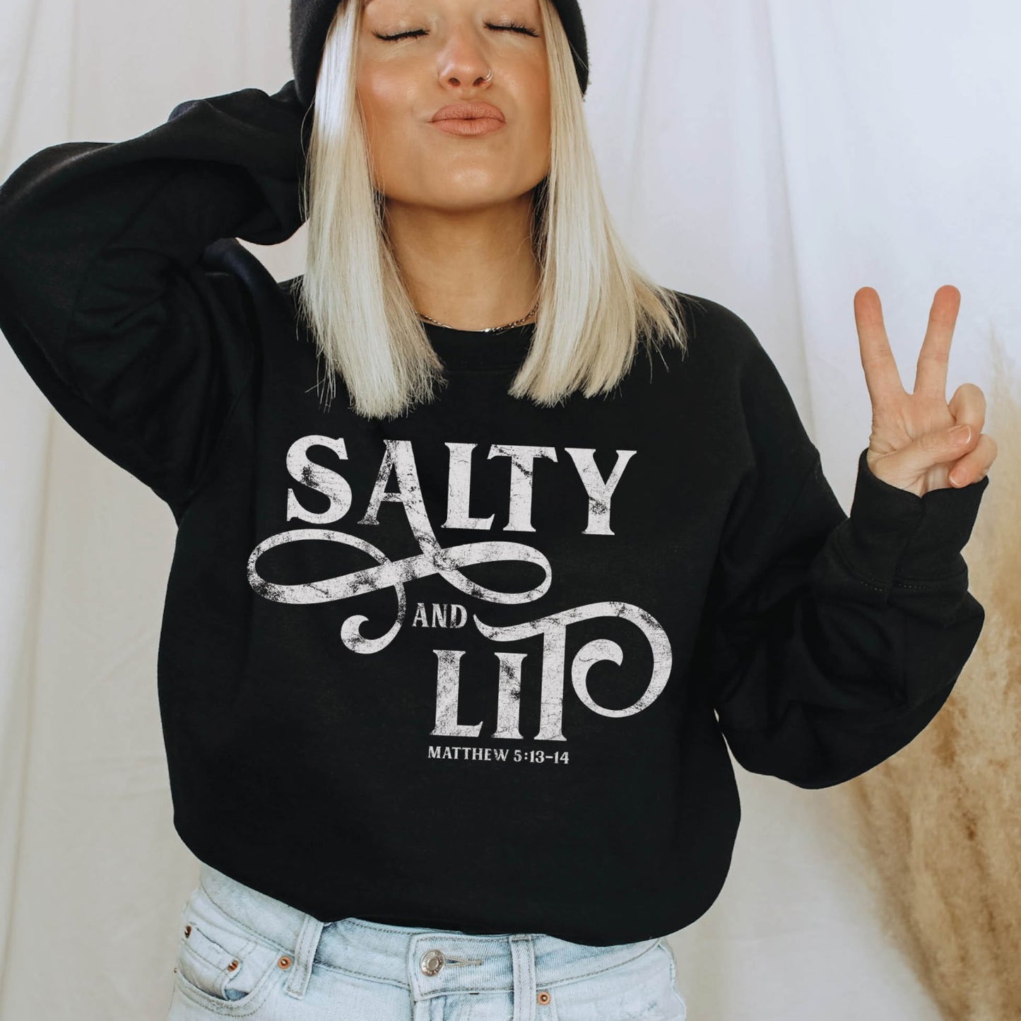 Young trendy woman wearing a faith-based Salty And Lit Matthew 5:13-14 bible verse funny Christian aesthetic unisex crewneck sweatshirt with distressed swirl typography design printed in white on cozy black sweater for women