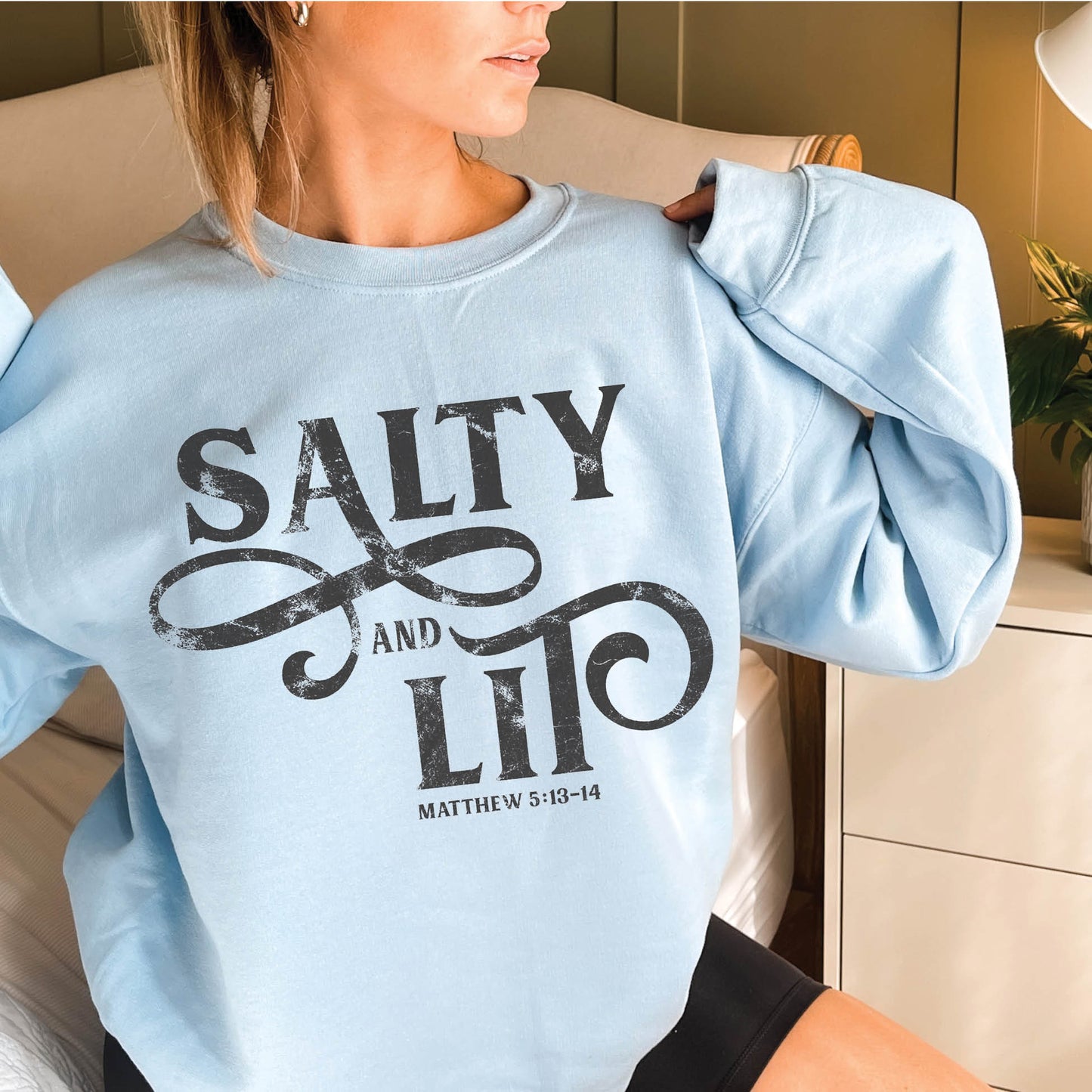 Woman wearing an oversized faith-based Salty And Lit Matthew 5:13-14 bible verse funny Christian aesthetic unisex crewneck sweatshirt with distressed swirl typography design printed in charcoal gray on cozy light blue sweater for women