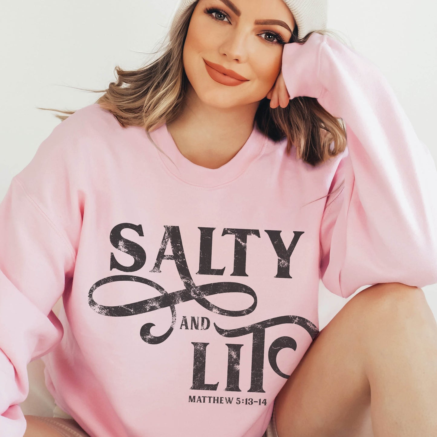 Trendy young woman wearing an oversized faith-based Salty And Lit Matthew 5:13-14 bible verse funny Christian aesthetic unisex crewneck sweatshirt with distressed swirl typography design printed in charcoal gray on cozy pink sweater for women