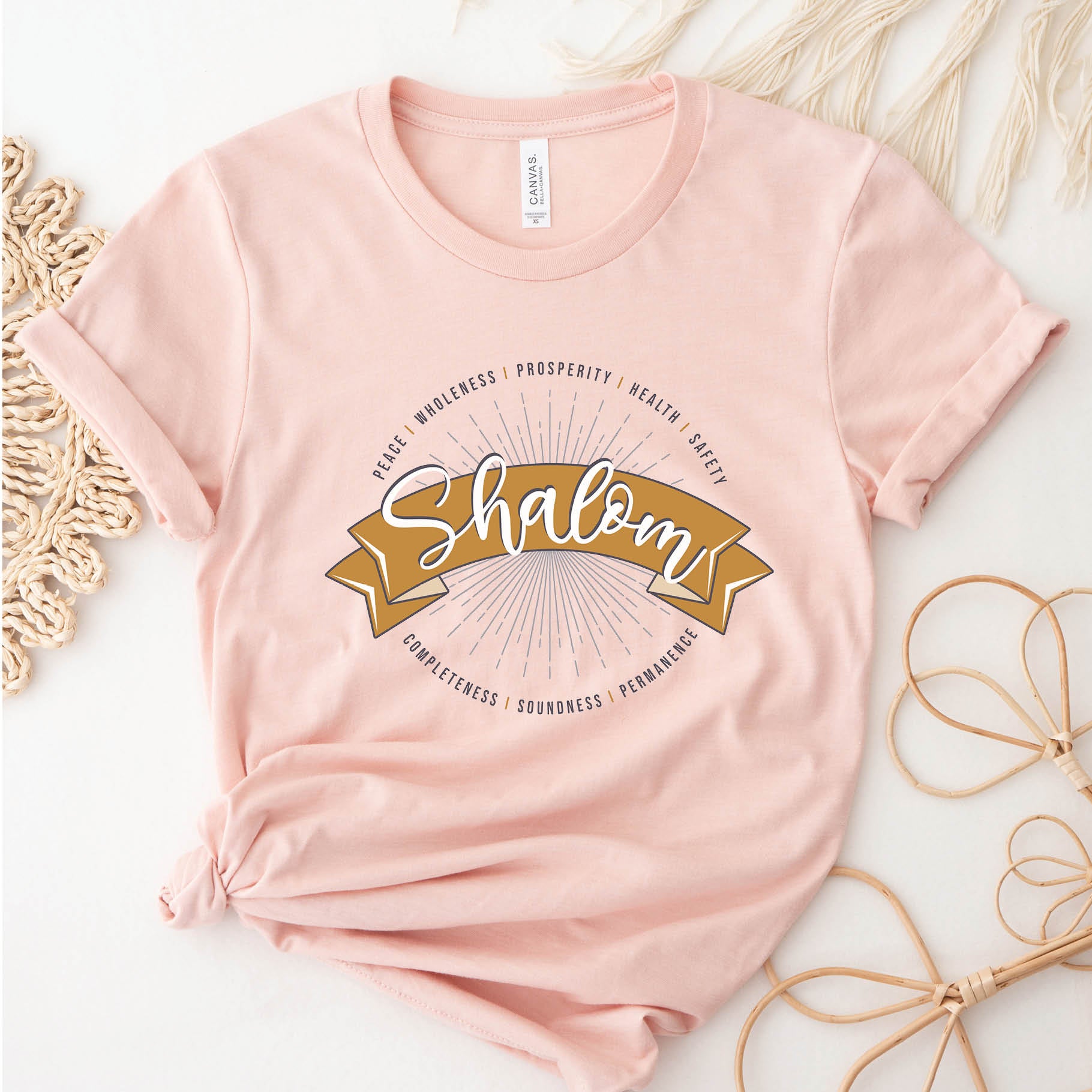 Soft heather prism peach faith-based Christian women's unisex t-shirt with a gold banner and sunburst design that says the Biblical Hebrew meaning of the word "SHALOM" - Peace, Completeness, Prosperity, Health, Safety, Wholeness, Soundness, and Permanence definition, makes a great mother's day gift for mom!