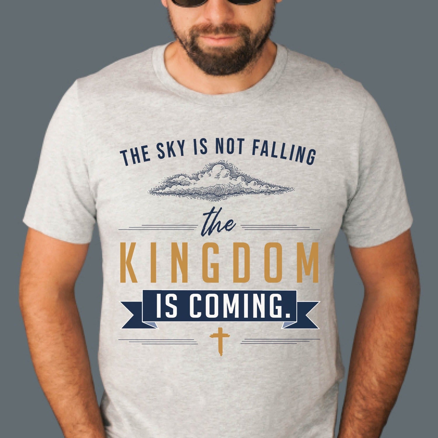 Man, husband, or Dad wearing a heather sport gray unisex t-shirt with a Christian Kingdom of God through Jesus faith-based message that says, "The Sky Is Not Falling, The Kingdom is Coming" with bold font, vintage clouds, banner, and cross design in navy blue and gold, created for bible study Men and Women believers, 2024 eclipse