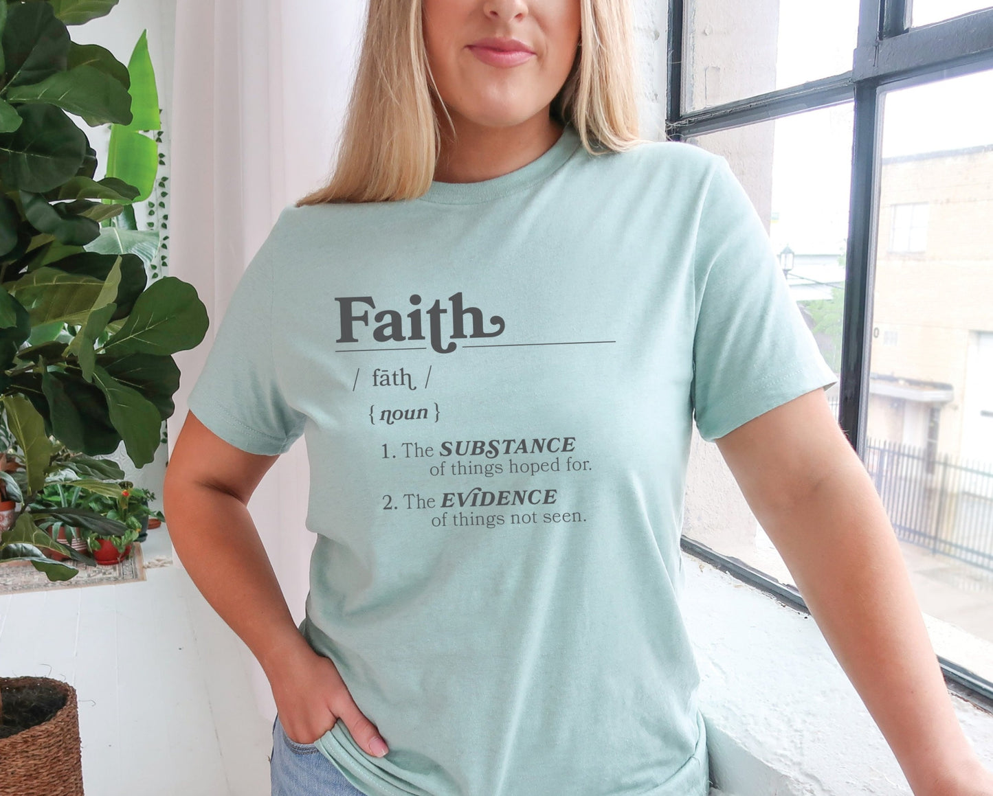 Young woman wearing a Faith Definition Hebrews 11:1 Christian aesthetic design printed in charcoal gray on soft heather dusty blue unisex t-shirt for women and men