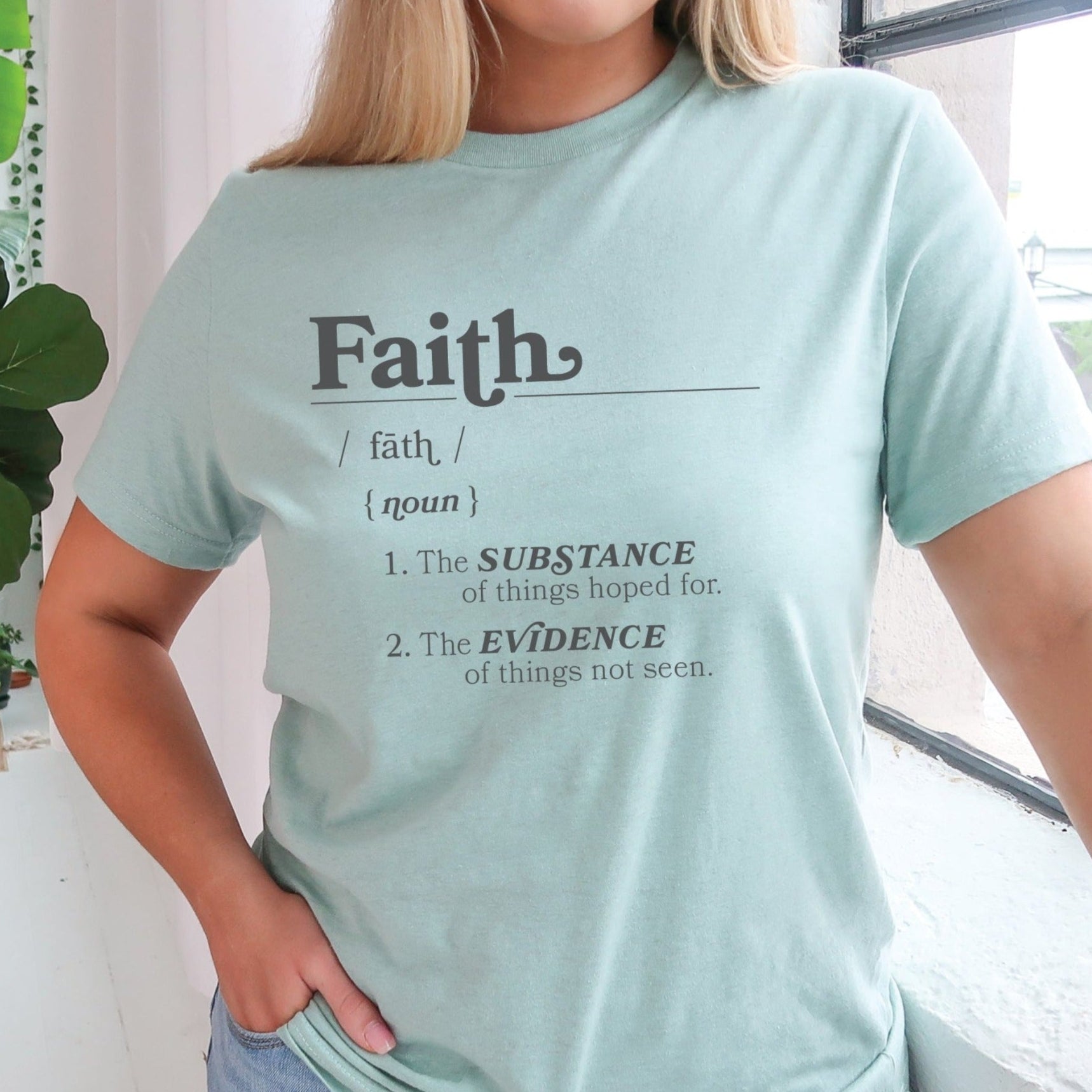 Young woman wearing a Faith Definition Hebrews 11:1 Christian aesthetic design printed in charcoal gray on soft heather dusty blue unisex t-shirt for women and men