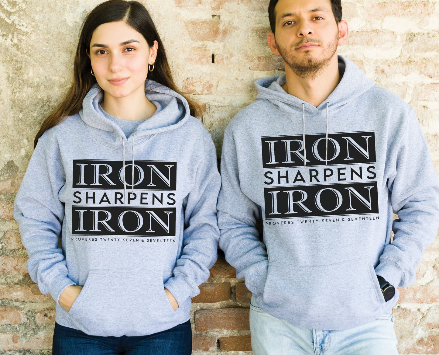 Young man and woman couple wearing Iron Sharpens Iron Proverbs 27:17 Bible Verse Christian aesthetic faith-based hoodie with bold black design printed on cozy heather sport gray unisex hoodie sweatshirt for men and women