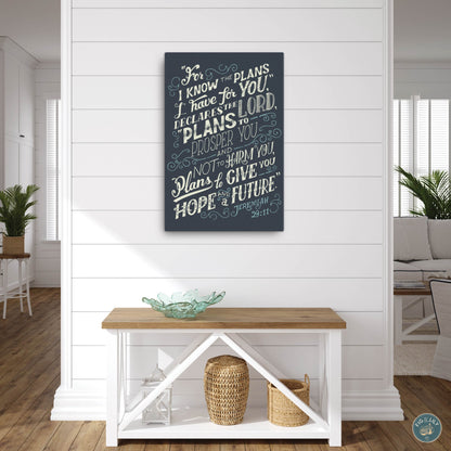 A 18x24" canvas wall art print featuring the text of Jeremiah 29:11 Bible verse in  cream and teal color typography with a moody navy blue background for home, church, or office decor, presented hanging on a white paneled farmhouse style modern home entrance hall wall. 