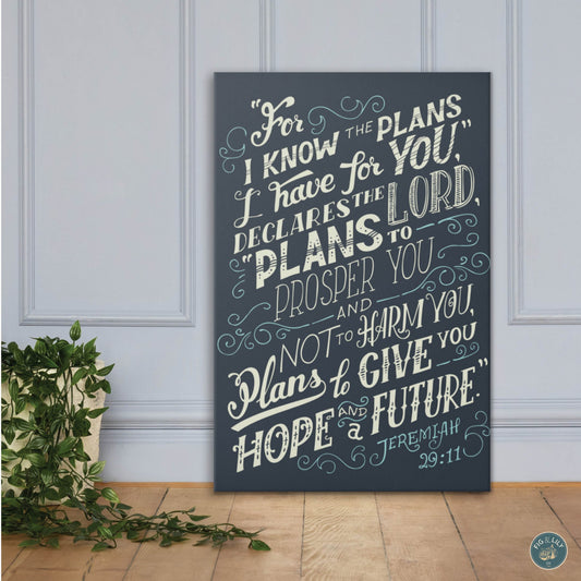 A 24x36" canvas wall art print featuring the text of Jeremiah 29:11 Bible verse in a cream color gradient background with navy blue background for home, church, or office decor, presented propped against a beautiful smoky blue paneled wall on a wooden floor. 
