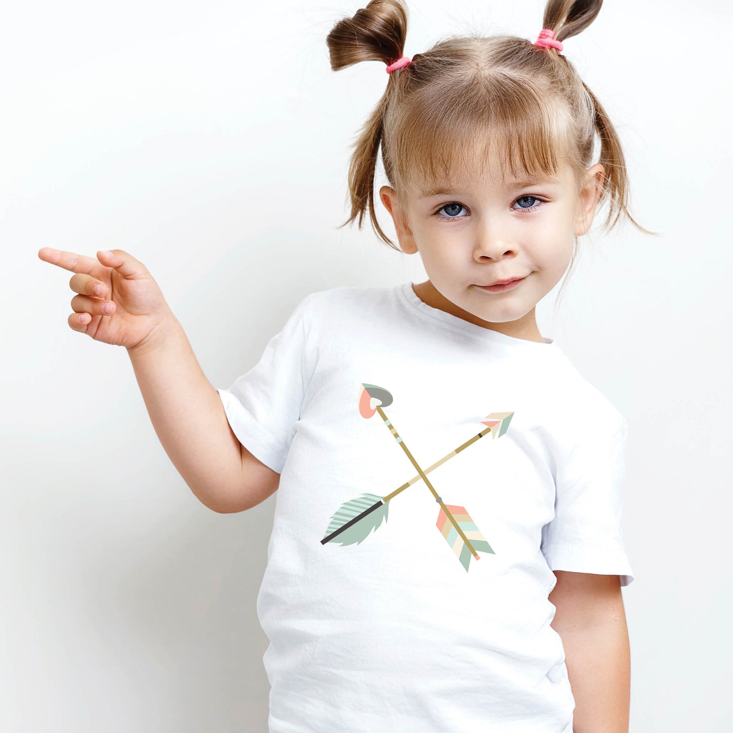 Funny little girl wearing a white toddler size t-shirt with teal blue and dusty pink pastel criss-cross boho arrows, one with a heart on the end, to match Christian mom's womens' "Raising Arrows" Psalm 127:4-5 bible verse t-shirt for boys and girls