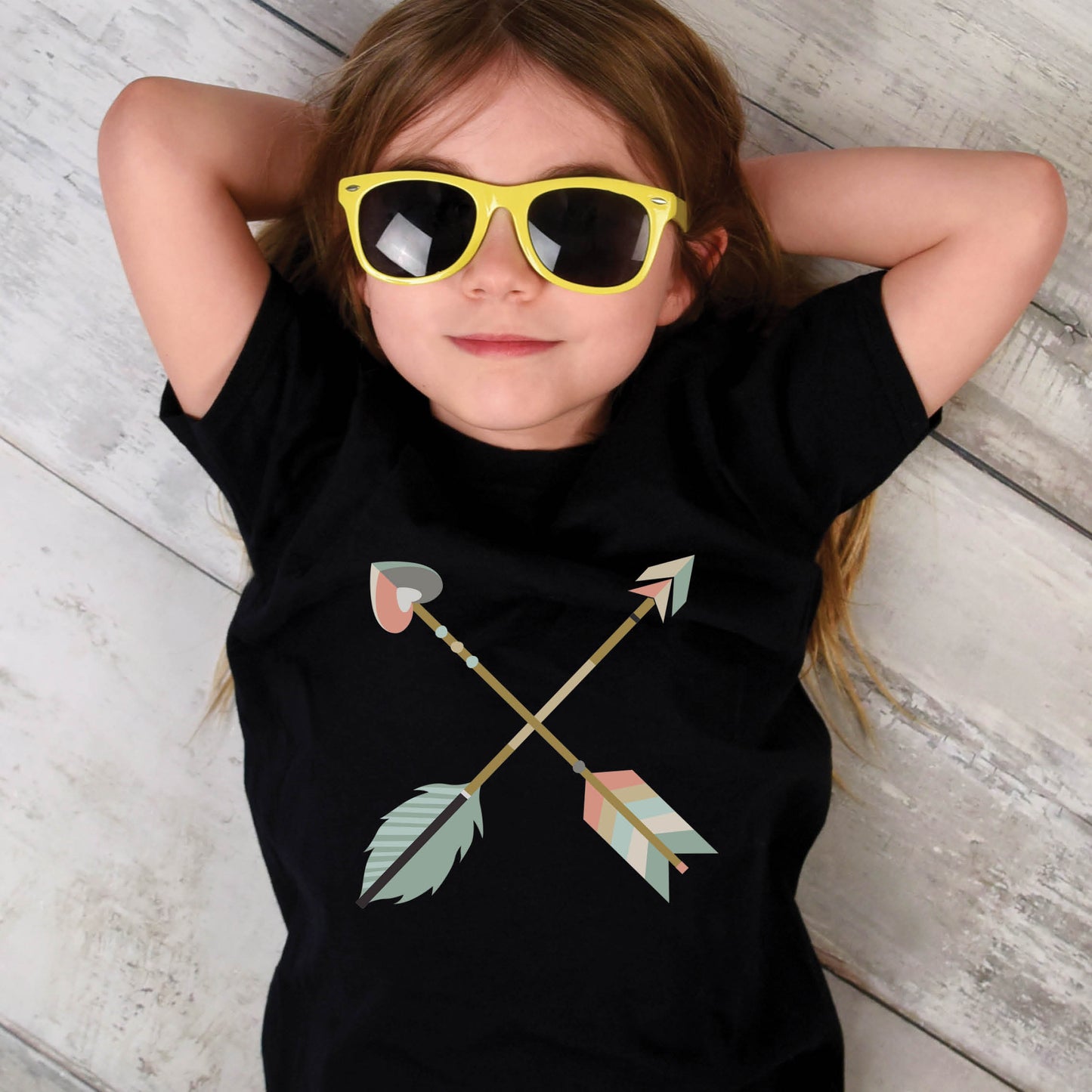 Funny young girl with yellow sunglasses wearing a black Youth size t-shirt with teal blue and dusty pink pastel criss-cross boho arrows, one with a heart on the end, matching mommy-and-me Christian Mom "Raising Arrows" Psalm 127:4-5 bible verse t-shirt for boys and girls