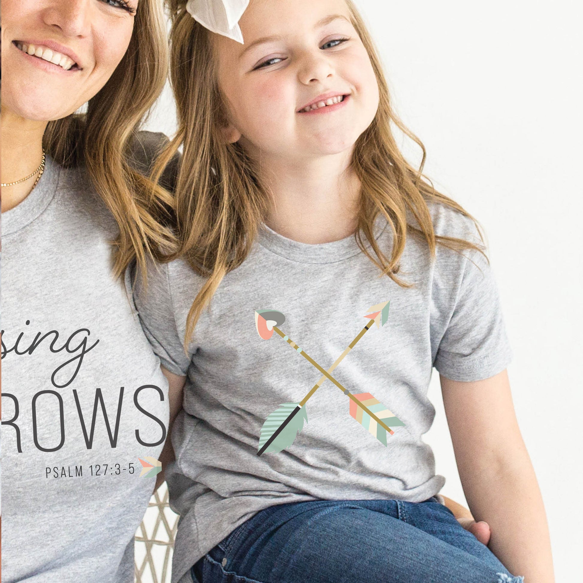 Young girl wearing a matching mommy-and-me Youth size t-shirt in heather gray with teal blue and dusty pink pastel criss-cross boho arrows, one with a heart on the end, to match Christian homeschool mom's / women's "Raising Arrows" Psalm 127:4-5 bible verse t-shirt for boys and girls