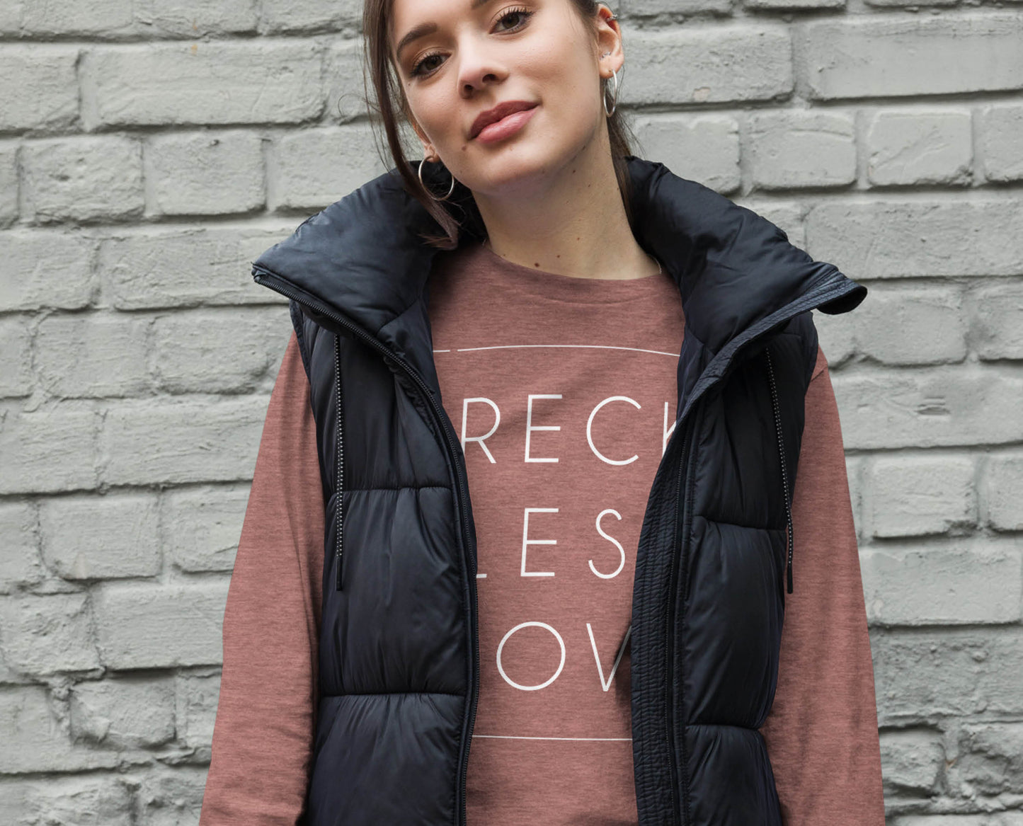 Young woman wearing puffer vest and a Rectangle Reckless Love Christian aesthetic worship design printed in white on cozy mauve dusty rose unisex long sleeve tee shirt for men and women